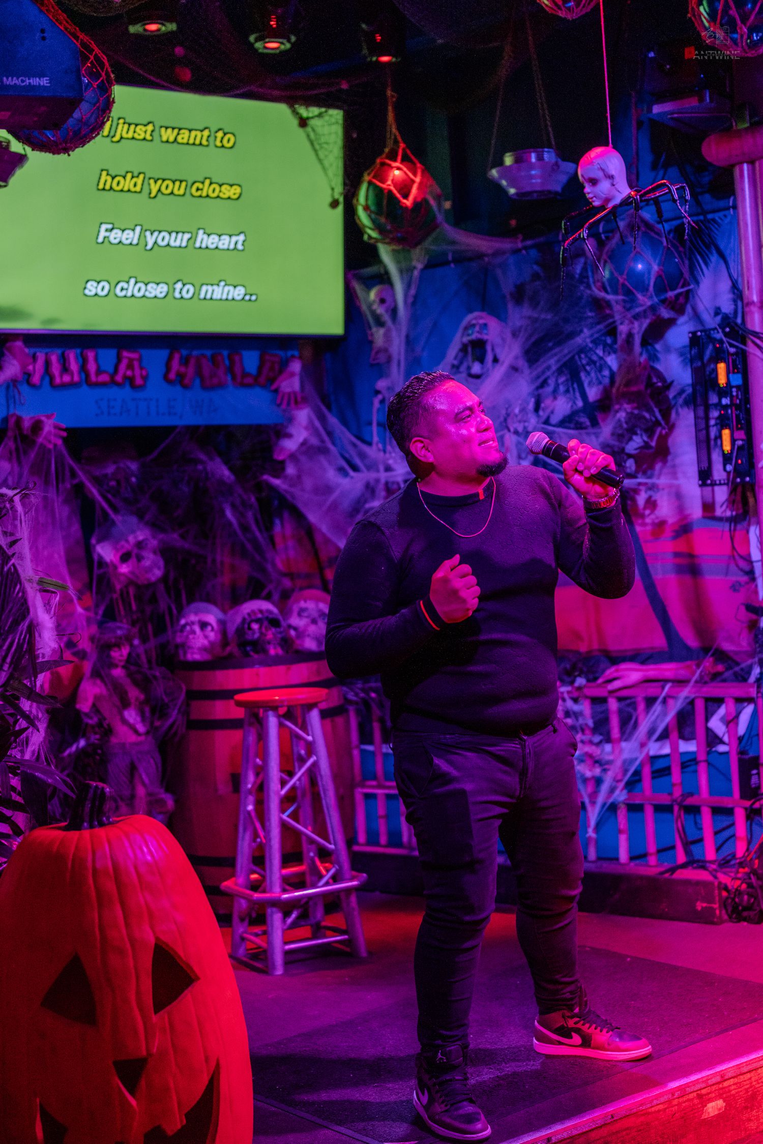 Person singing karaoke on Halloween-decorated stage with pumpkins, cobwebs, and spooky skulls under colorful neon lights.