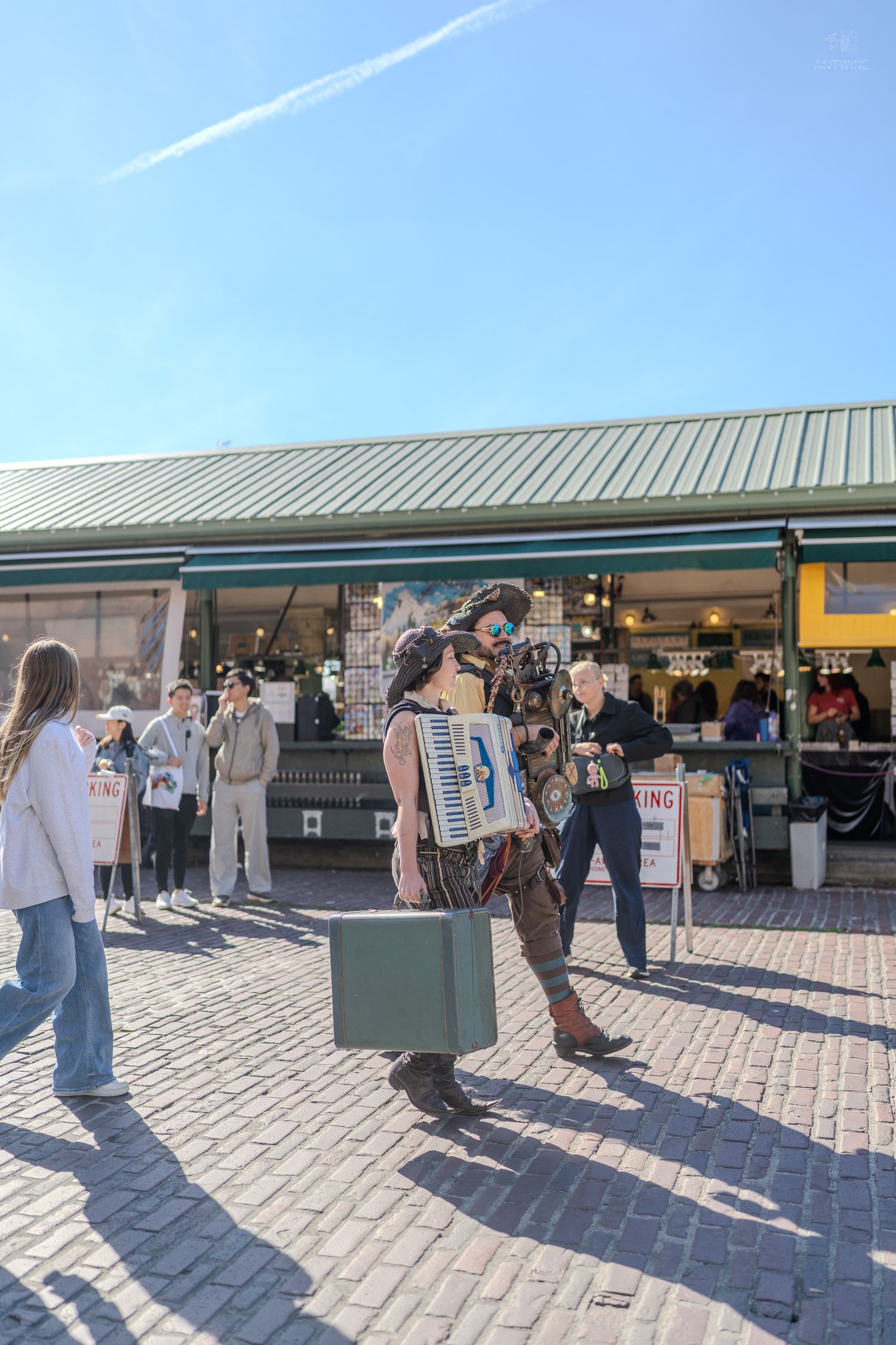 Street performer carrying an accordion and suitcase outside a market on a sunny day.