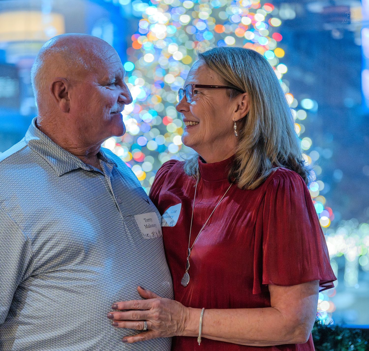 Couple smiling at each other at a festive Christmas party with colorful holiday lights in the background.