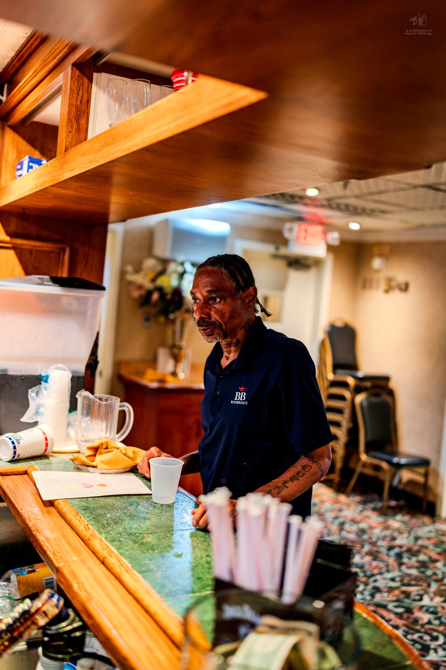 Bartender standing behind a wooden bar counter inside a riverboat lounge.
