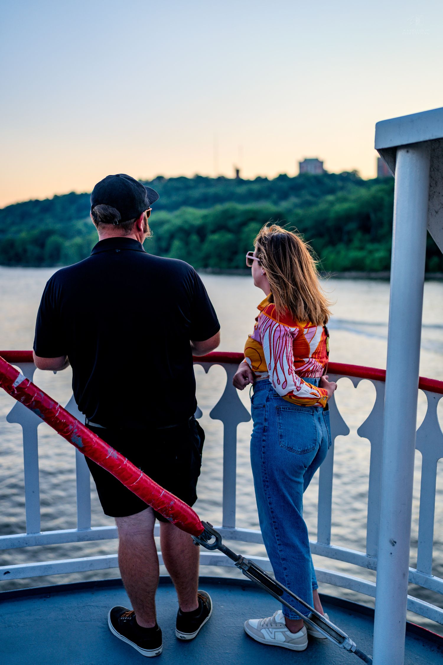 Couple standing at white and red railing on a boat deck enjoying a scenic river view at sunset.
