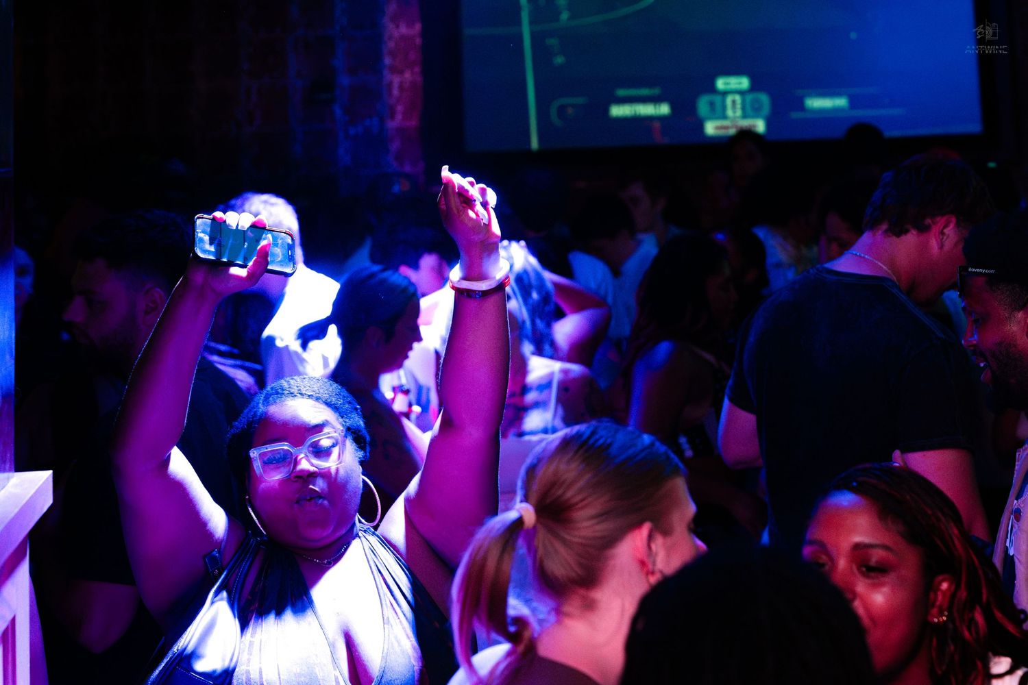 Woman dancing and recording video on phone under blue neon lights at a nightclub.