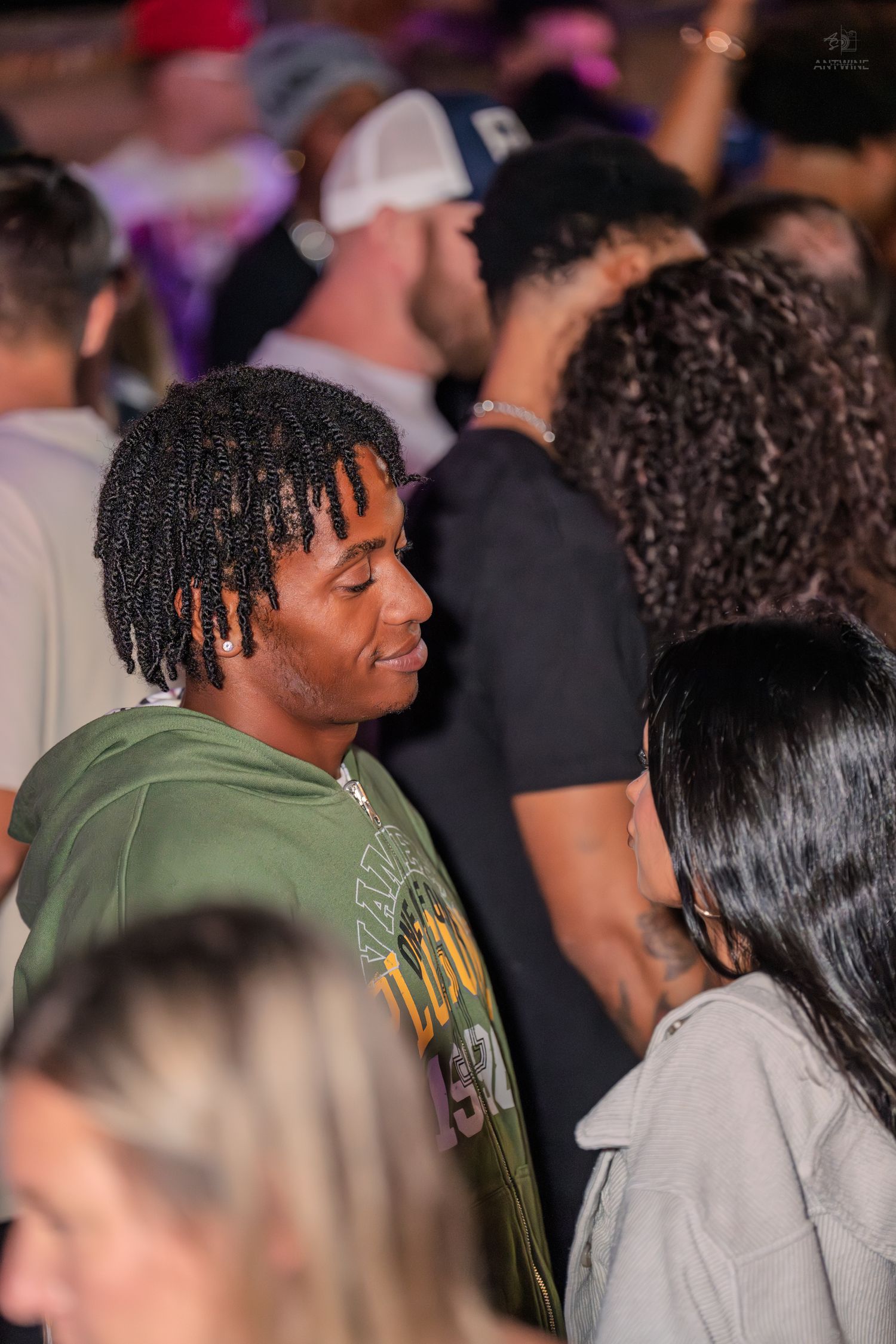 Young man in green hoodie with twist braids socializing at a busy nightclub event.