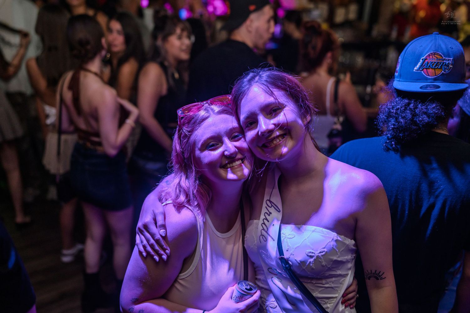 Two smiling young women pose together under purple lights at a lively nightclub party.