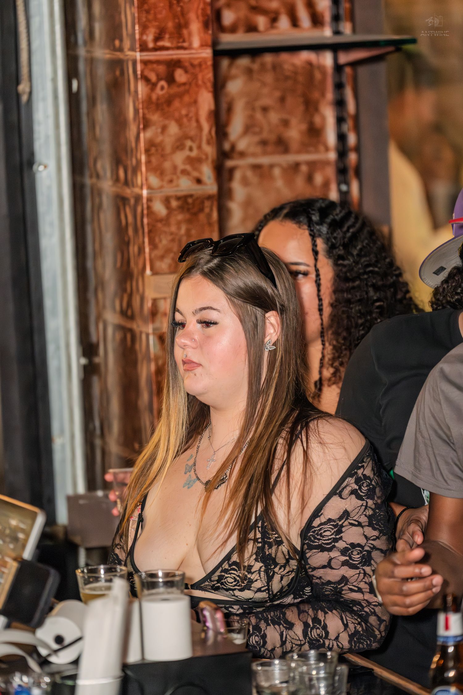 Woman in black lace top sits at a bar counter with others socializing in the dimly lit venue behind her.