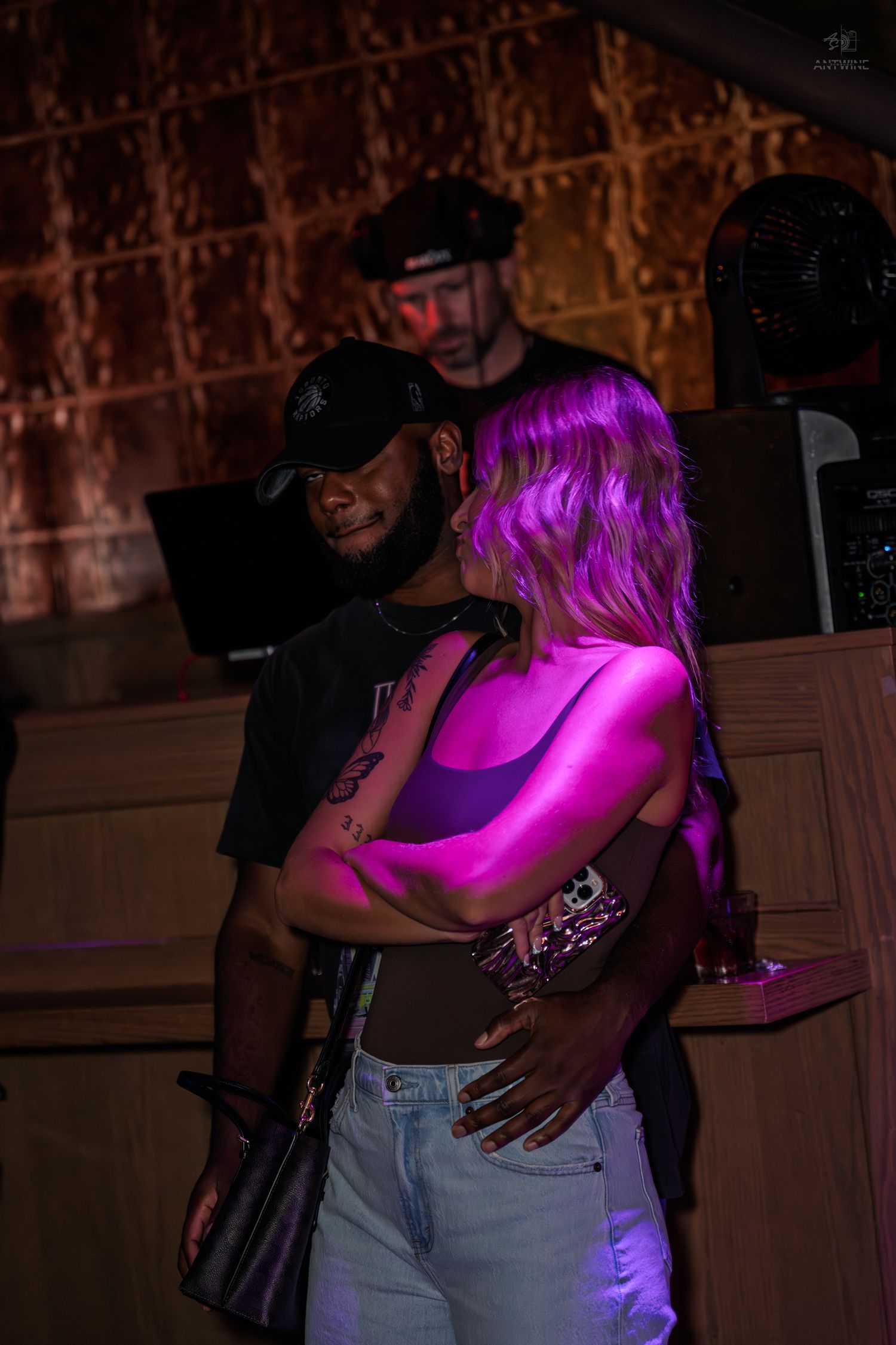 Couple dancing closely under pink neon lights at a nightclub with a DJ in the background.