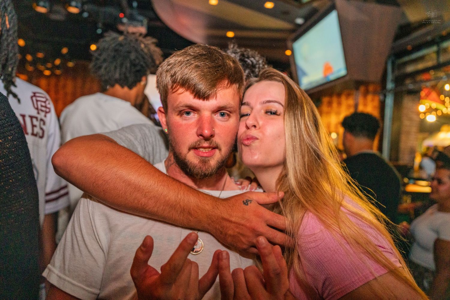 Young man and blonde woman posing closely together at a dimly lit bar at night.