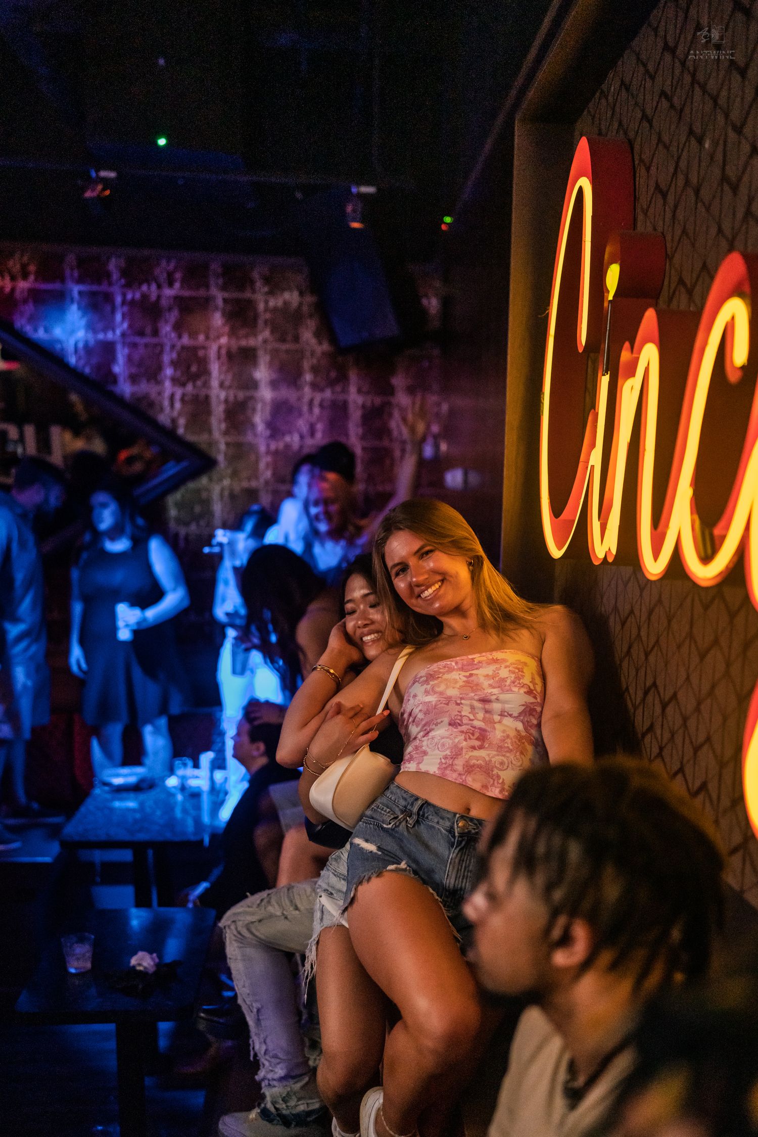 Two young women smiling and posing beside a glowing Cincy neon sign at a nightclub.