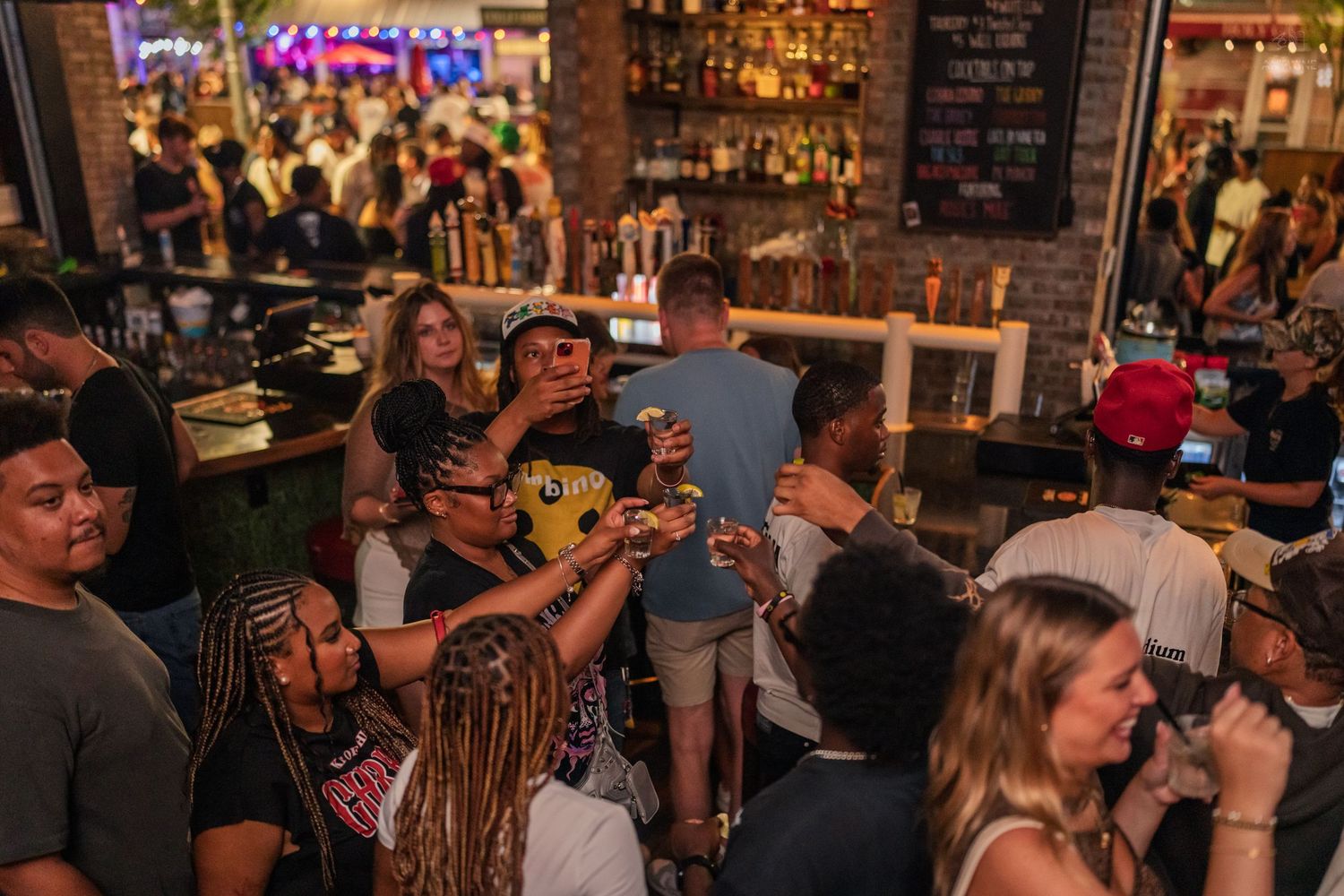 Lively crowd holding drinks and socializing at a busy nightclub bar at night.