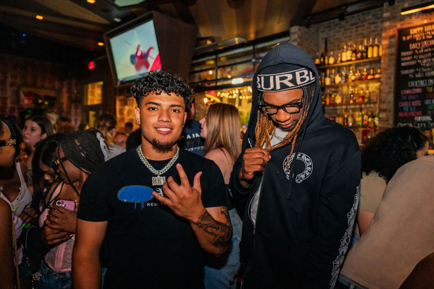 Two men posing together at a busy bar with bottle-lined shelves in background.