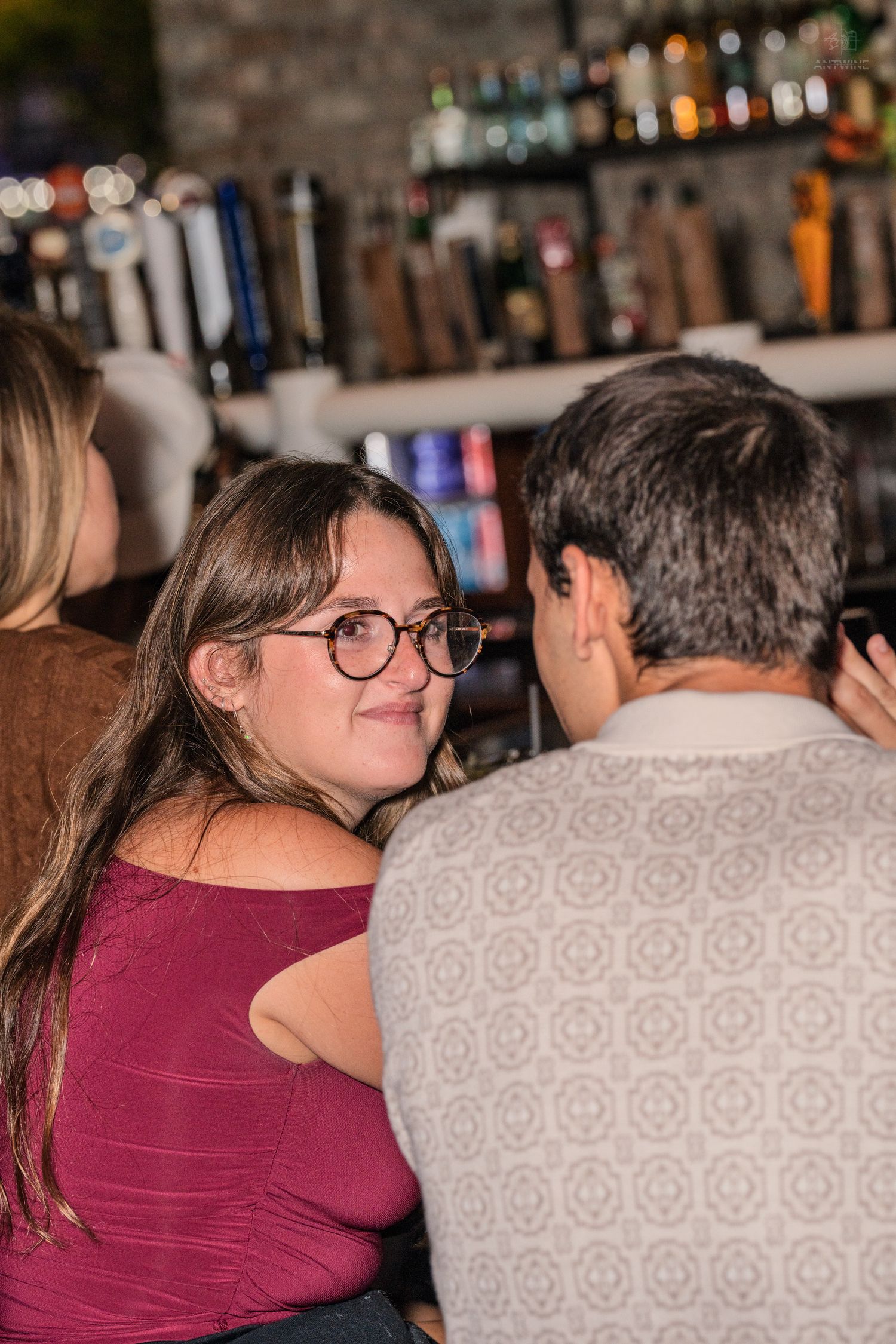 Smiling woman wearing round glasses in burgundy top glancing back at companion near a bar.
