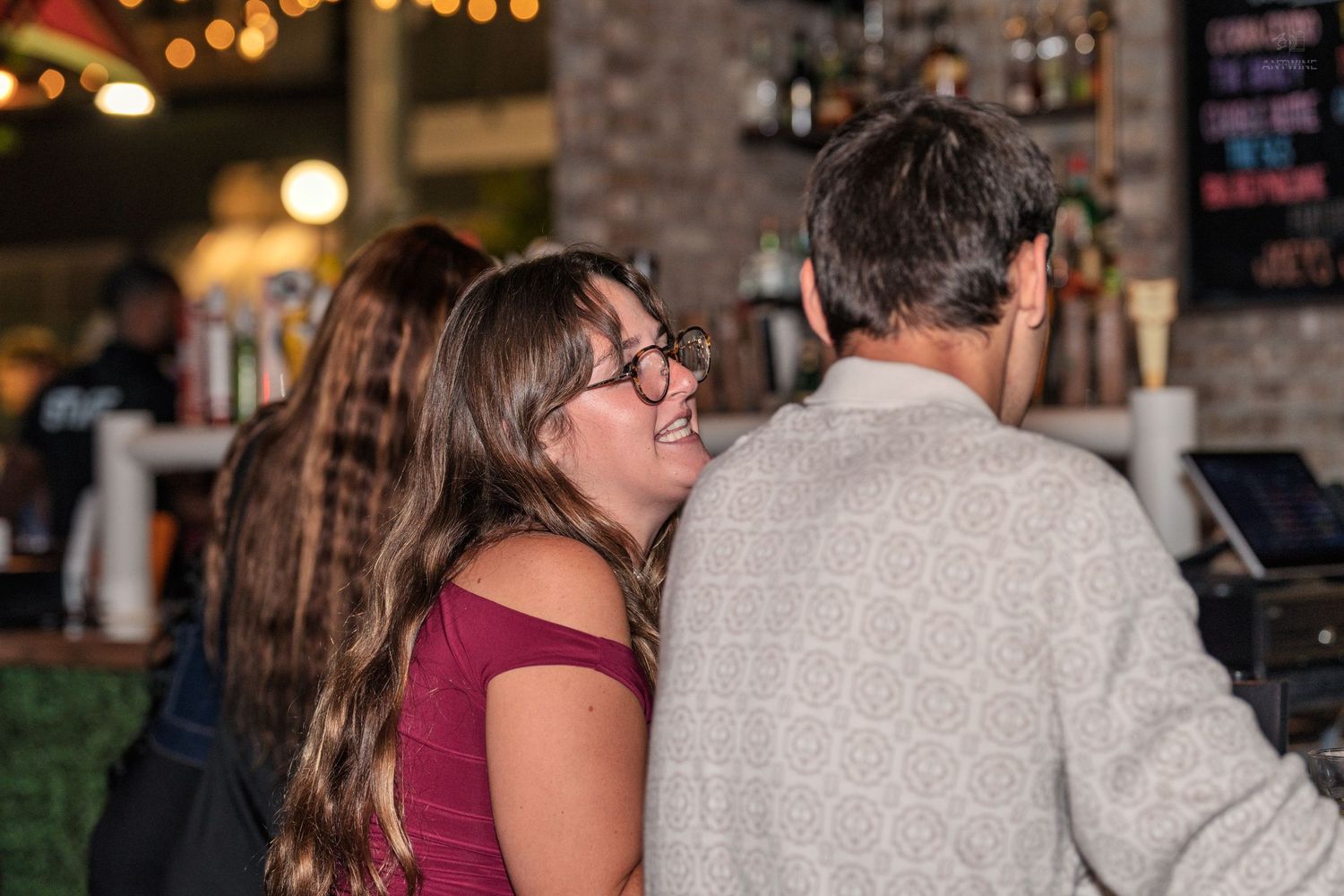 Woman with glasses smiling and laughing with a man at a bar counter inside a restaurant.