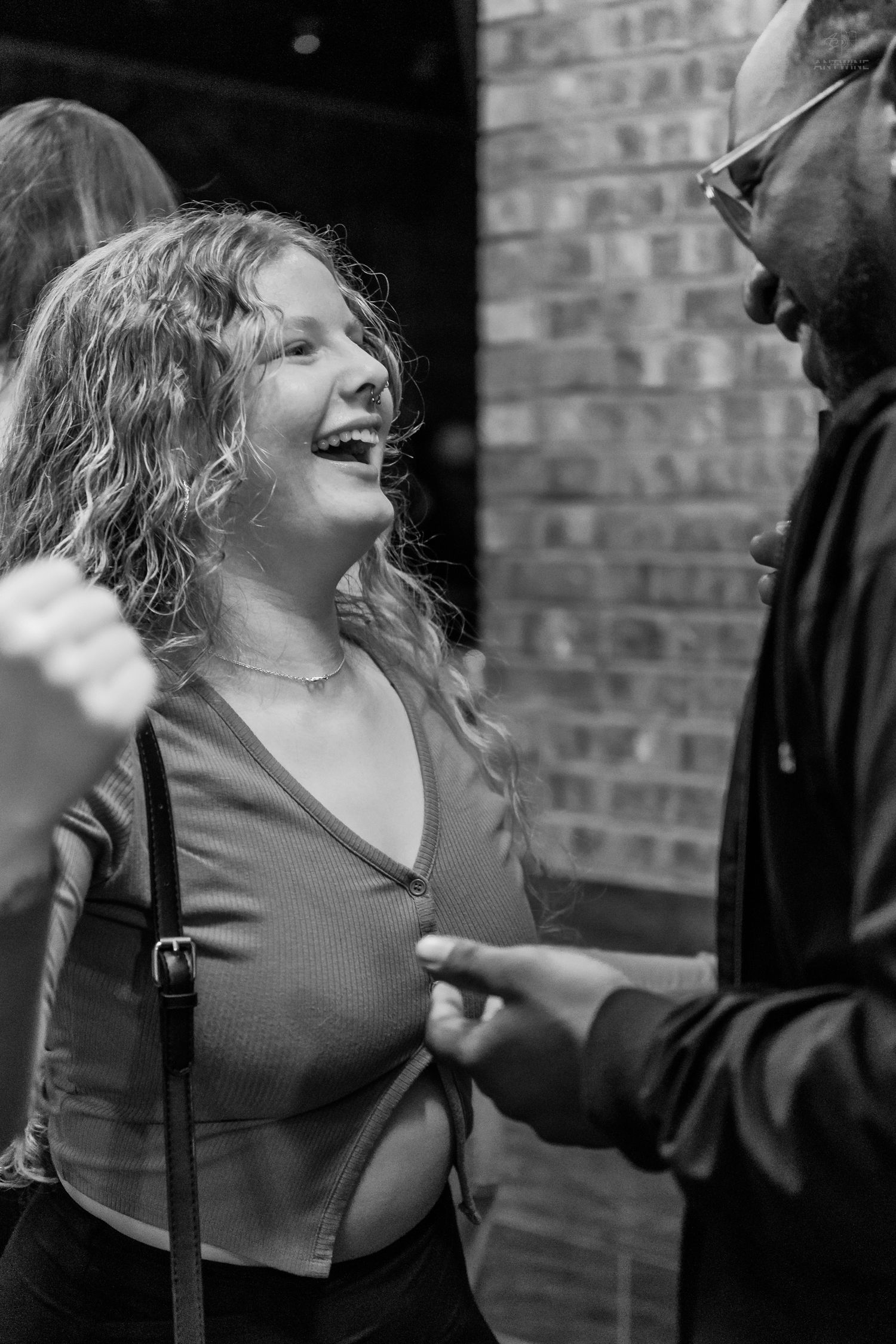 A curly-haired woman laughs joyfully while talking to a man outside a brick building.