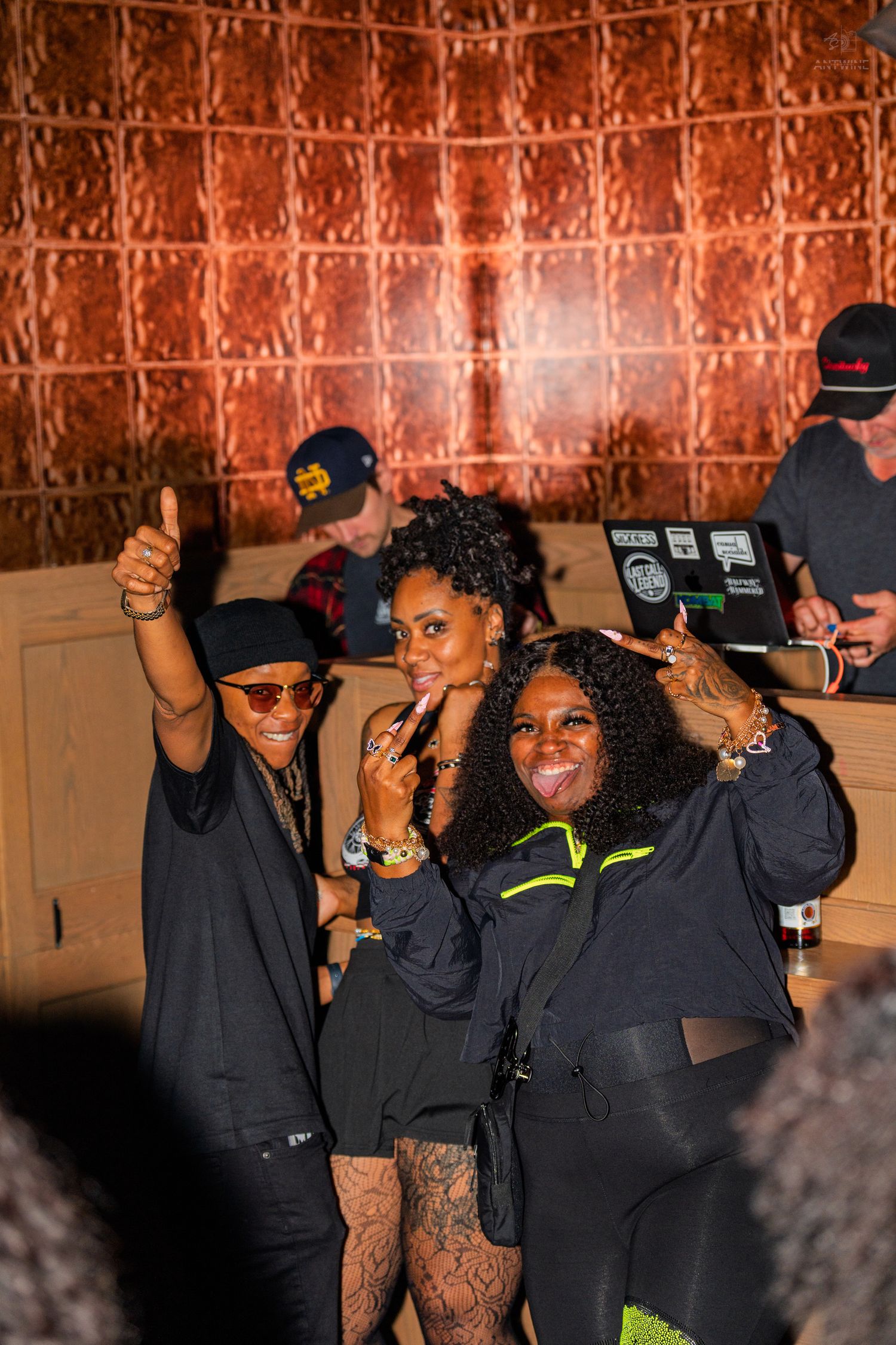 Three women posing and smiling near a DJ booth at a nightclub with copper-tiled walls.