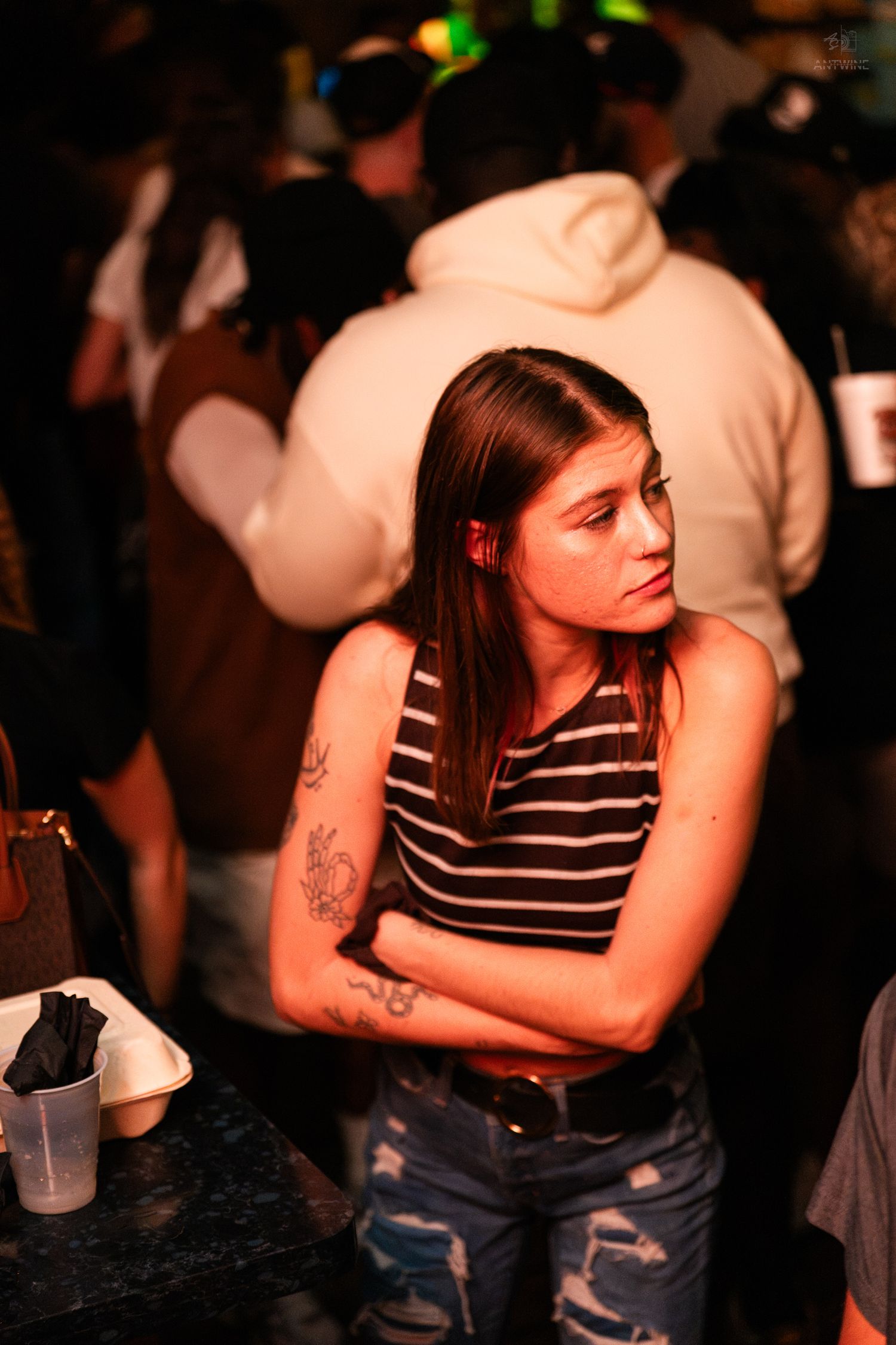 Young woman in striped tank top with tattoos standing at a crowded nightclub bar.