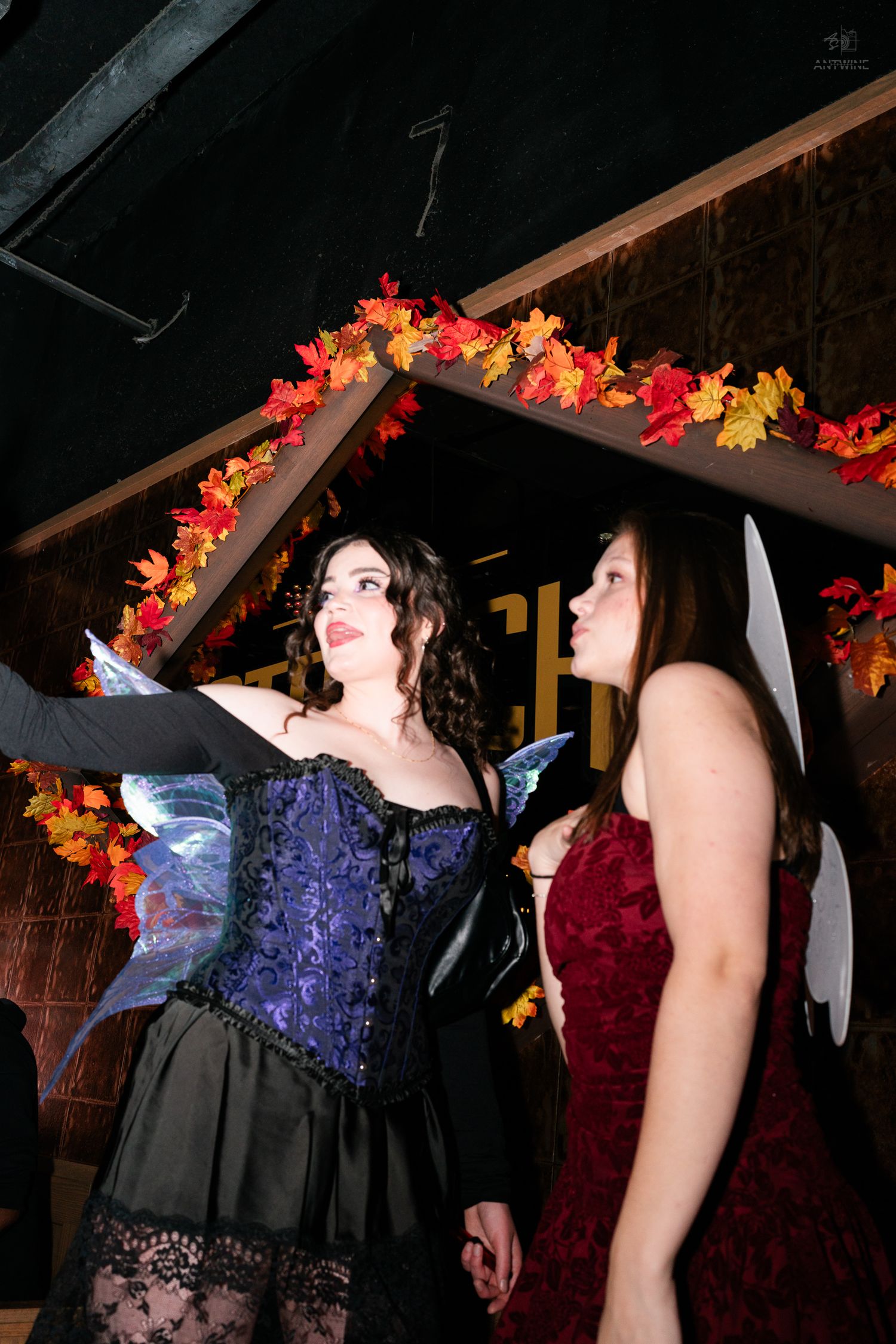 Two women in fairy Halloween costumes under autumn leaf decorations at a bar.