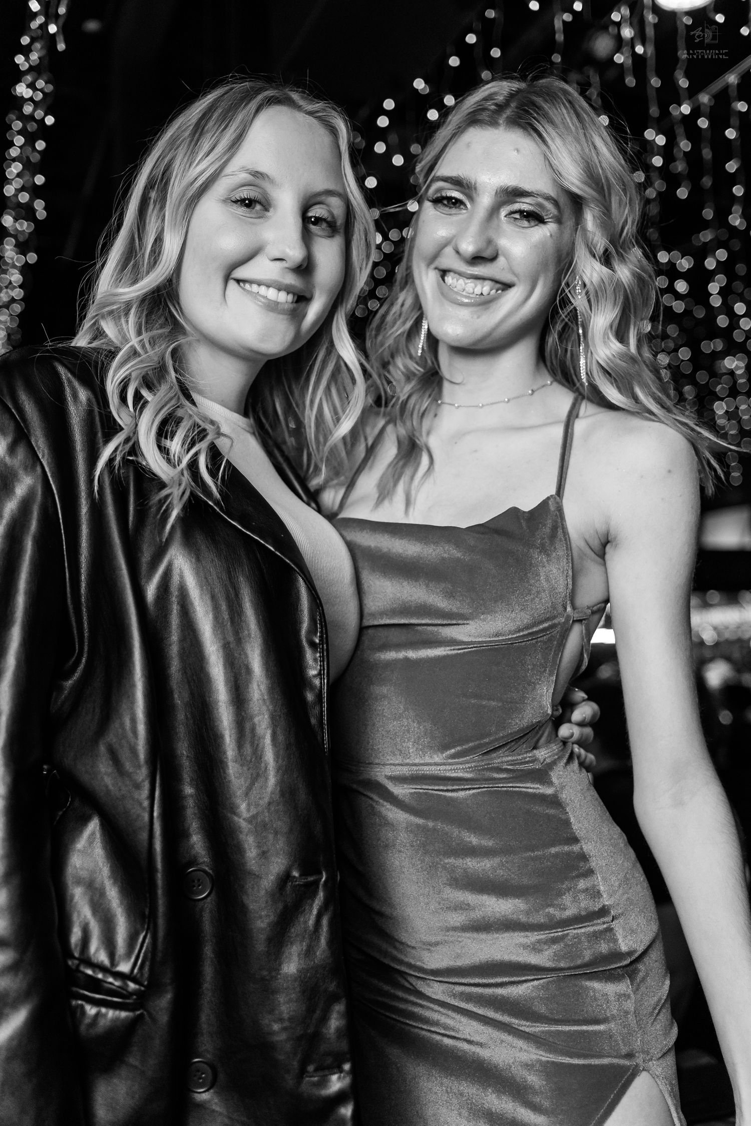 Two smiling blonde women pose together at a nightclub, one in a leather jacket and one in a satin dress.