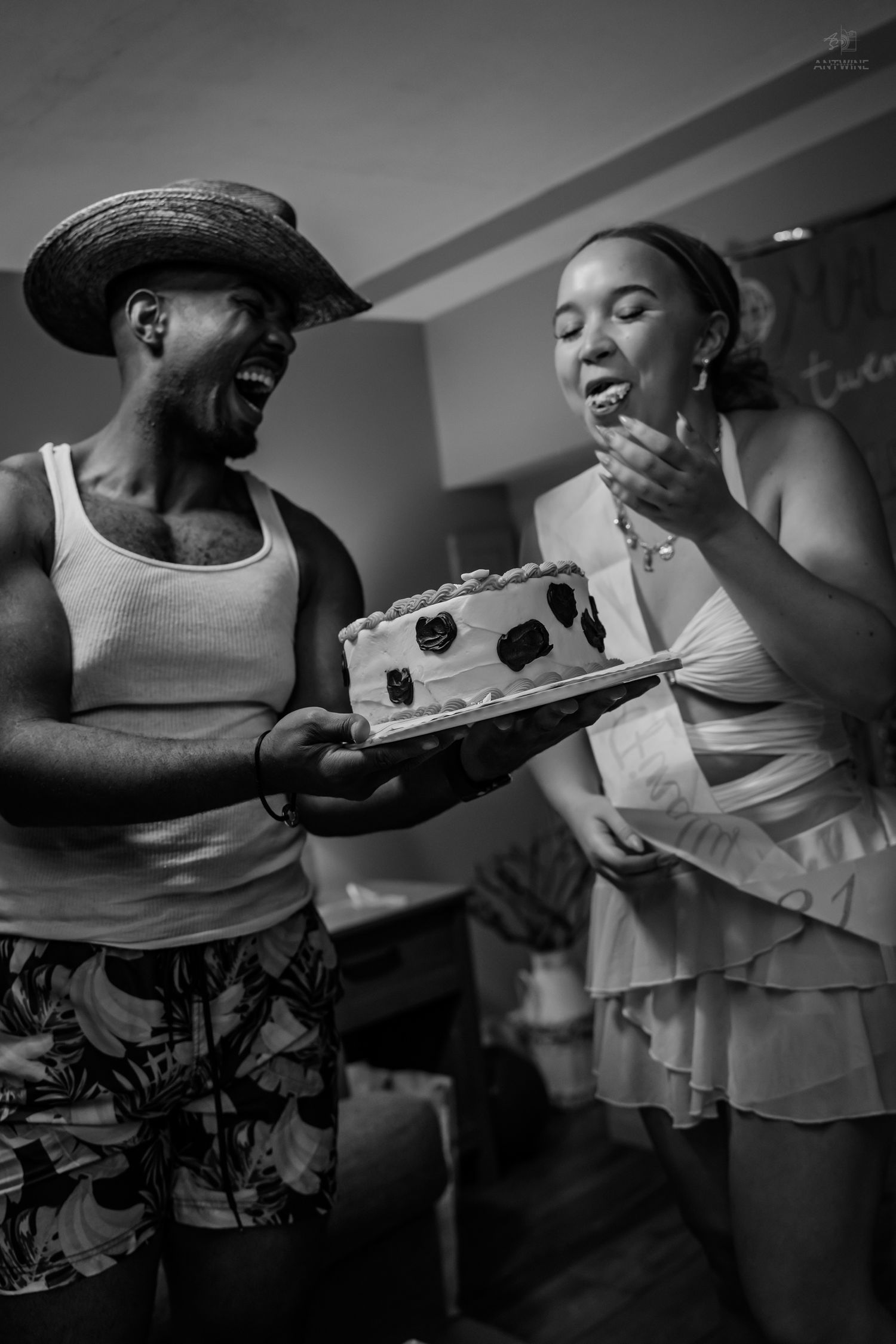 Black and white photo of man in straw hat holding cake as woman tastes frosting at birthday party.
