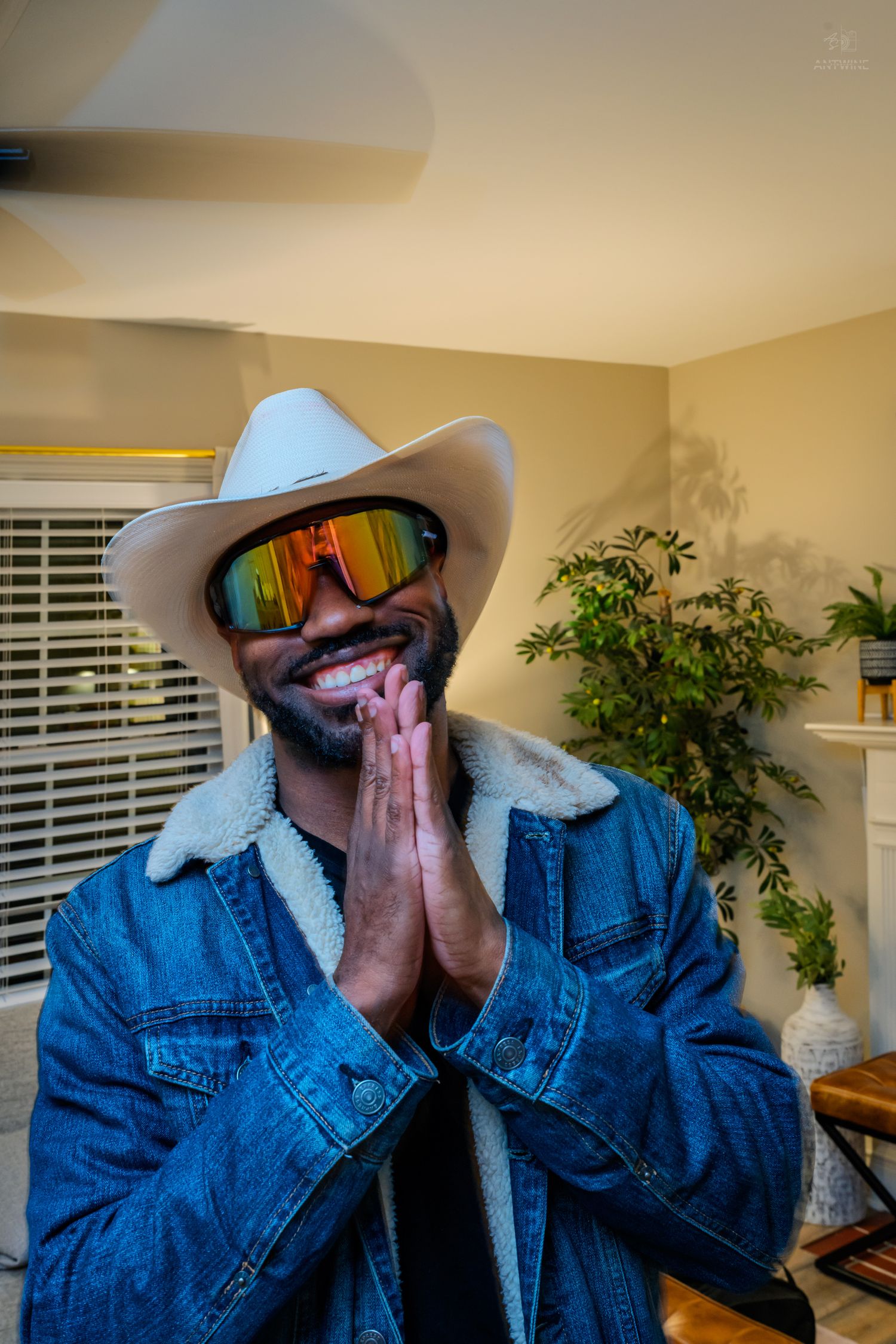 Smiling man in white cowboy hat and mirrored sunglasses wearing denim jacket with hands pressed together.