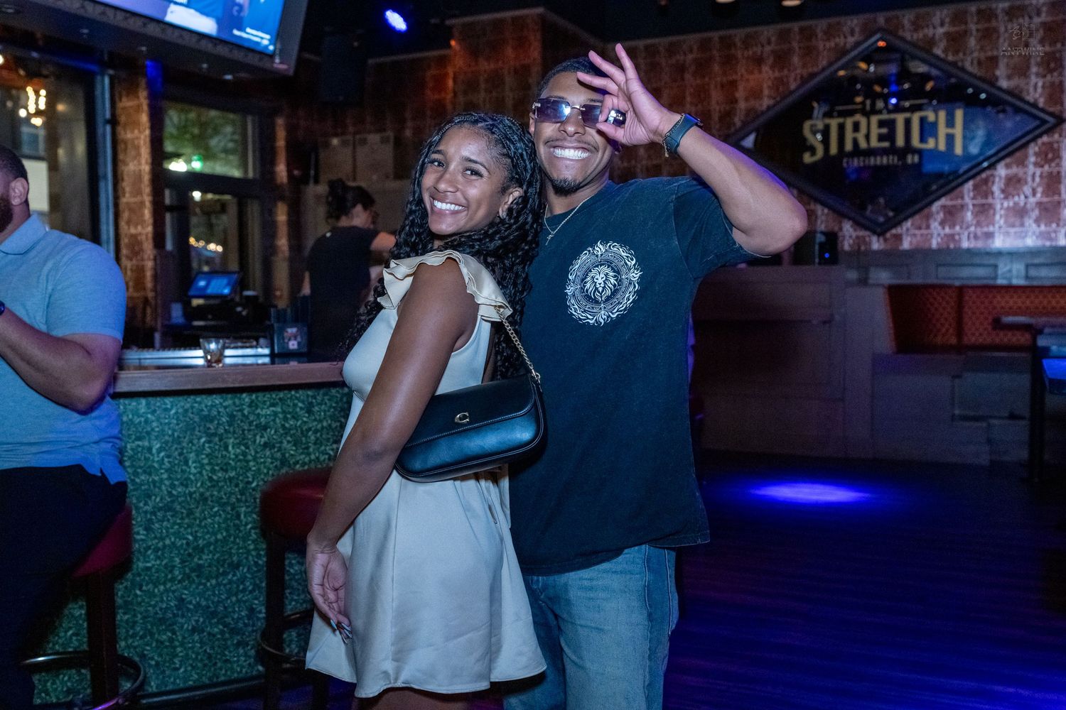 A happy couple poses together at a bar, with a Stretch sign glowing in the background.