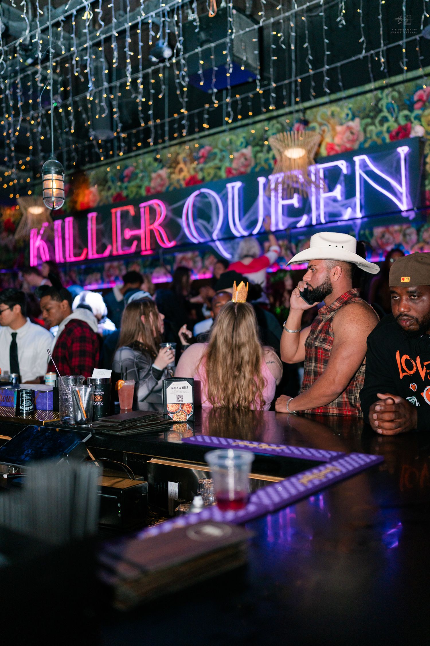 Patrons socializing at the Killer Queen bar featuring bright neon signage, string lights, and festive decor.