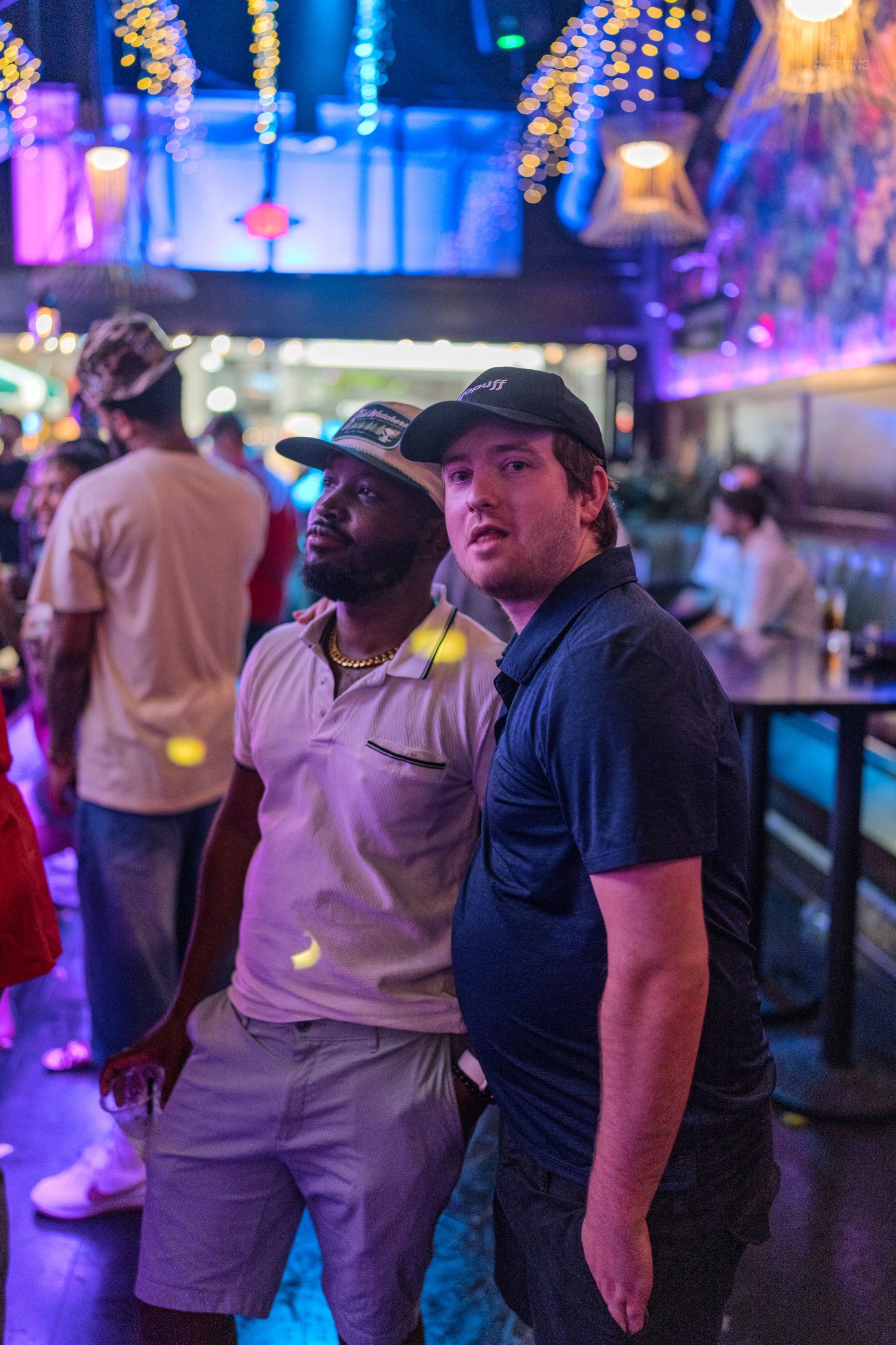 Two men posing together under colorful string lights inside a lively nightclub.