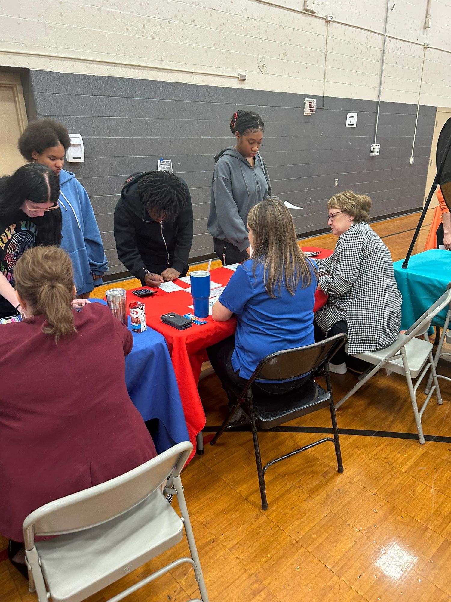 Students and teachers gather around a red-clothed table in a school gymnasium for an educational activity.