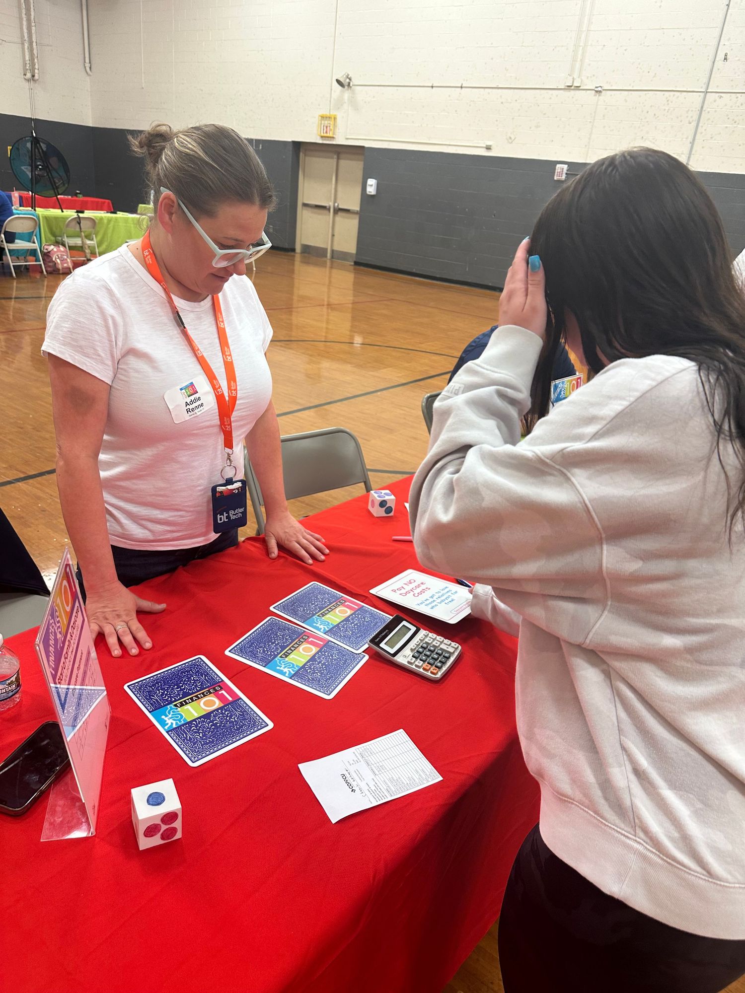 A woman in a white shirt presents educational card games at a red table in a school gym.