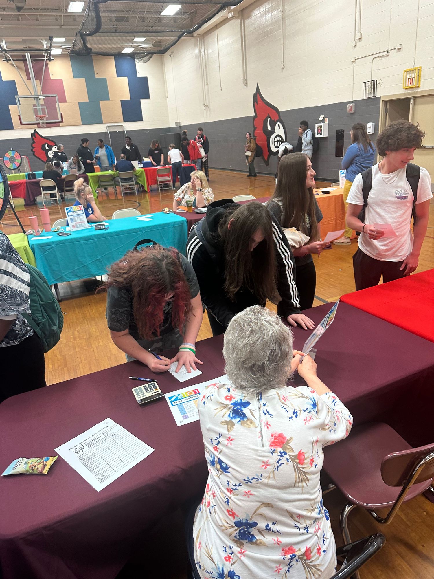 Students lean over a maroon table reviewing materials with an elderly representative at a school gymnasium career fair.