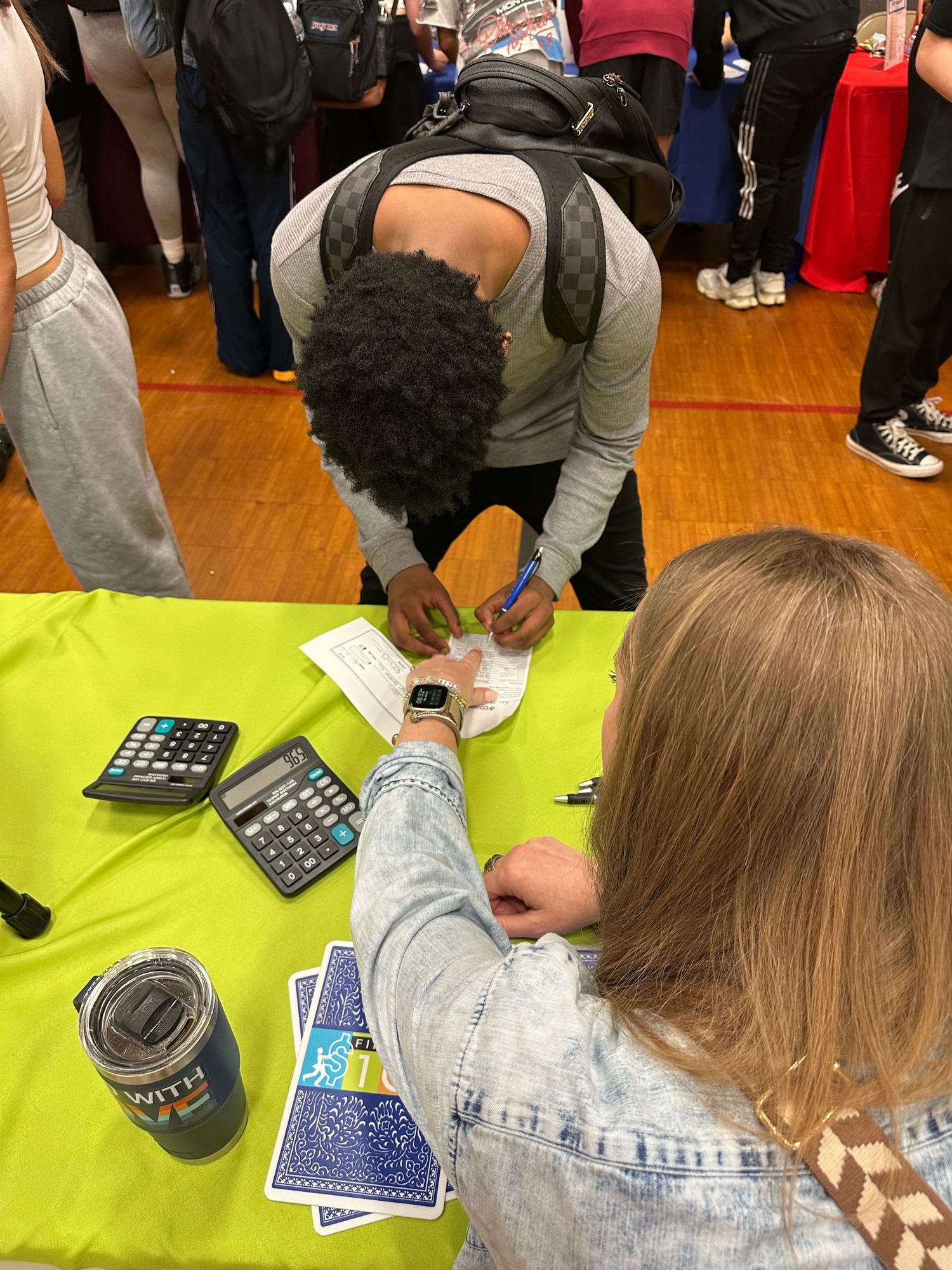 A student signs paperwork at a bright green table during a school career fair, with calculators and cards visible nearby.