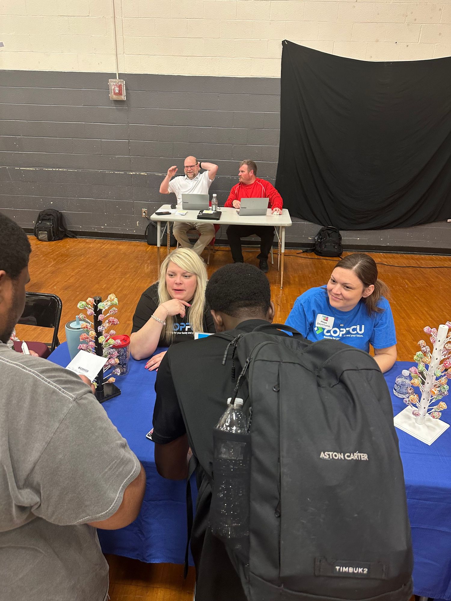 Students and representatives meet at a school career fair in a gymnasium, with a blue tablecloth and informational displays.
