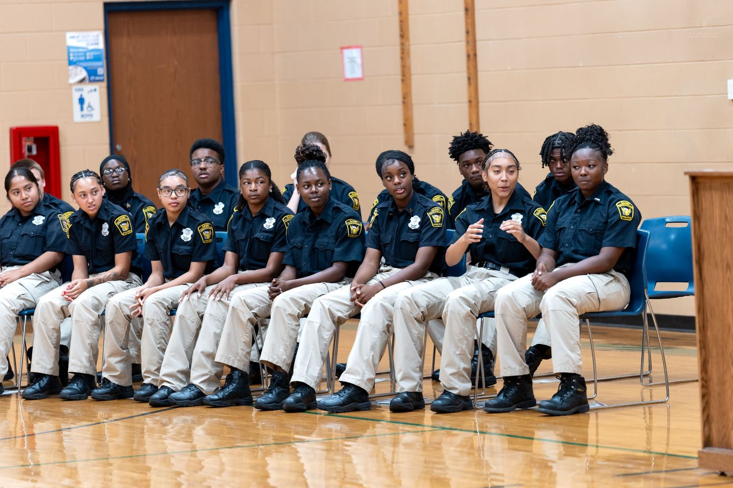 A group of young students in matching black uniforms and khaki pants sit in a row of chairs in a gymnasium.