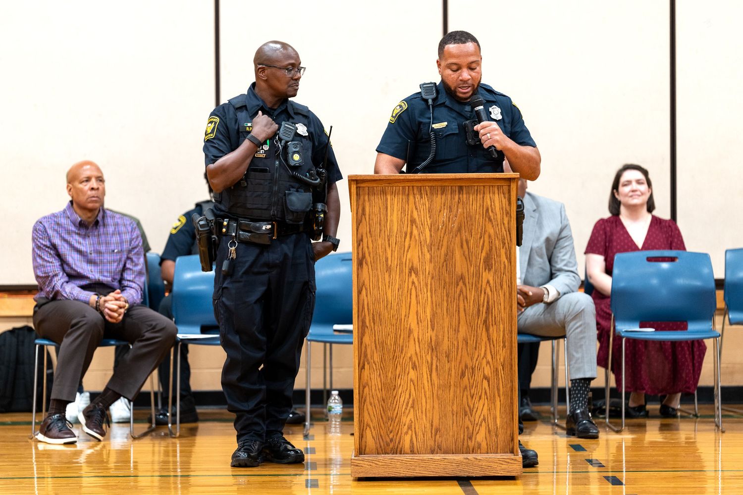 Two police officers speak at a podium during a community meeting in a gymnasium with officials seated behind them.