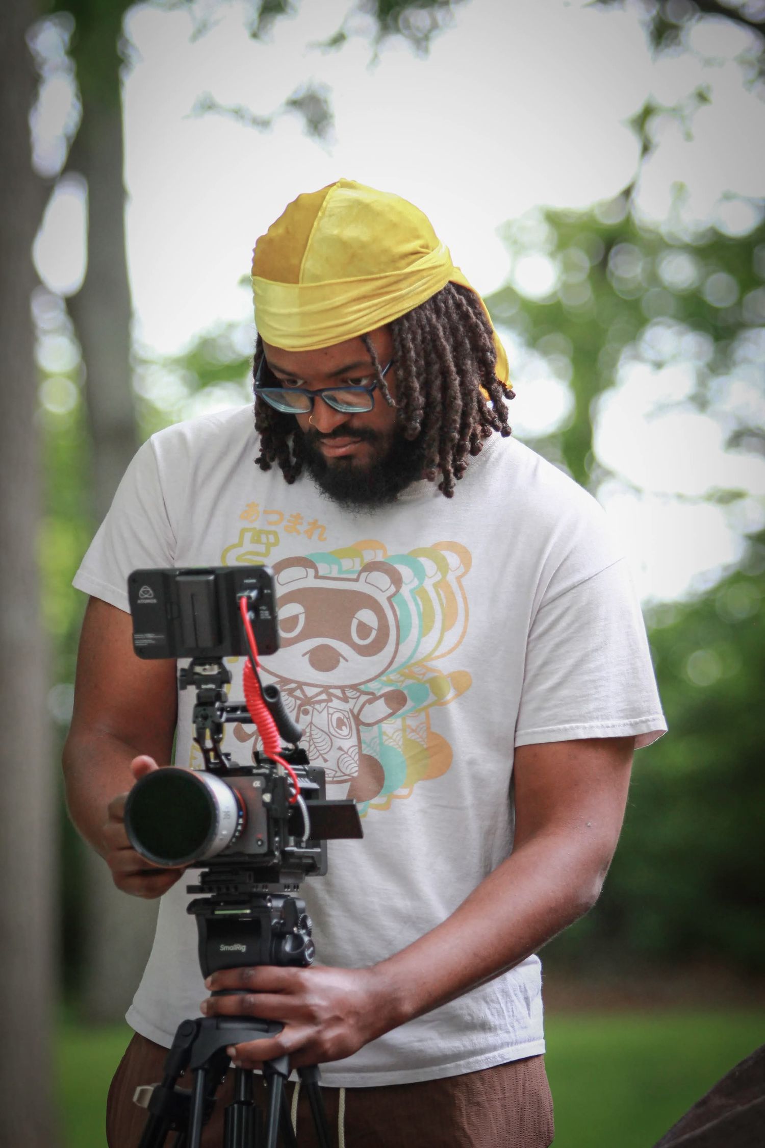 Videographer with dreadlocks and yellow hat operating a camera on a tripod outdoors.