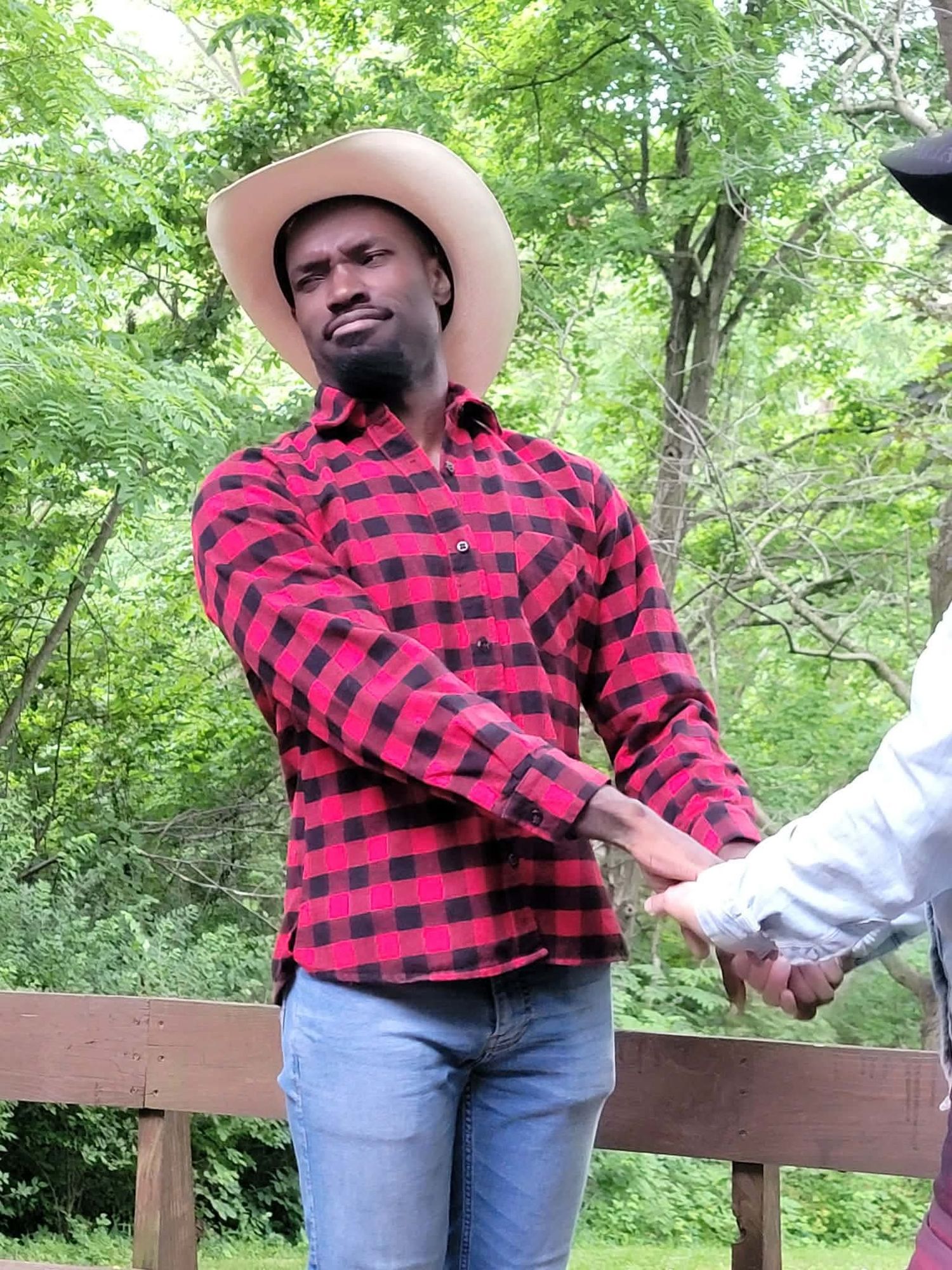 Man in red plaid flannel shirt and cowboy hat holding hands outdoors with green trees in background.