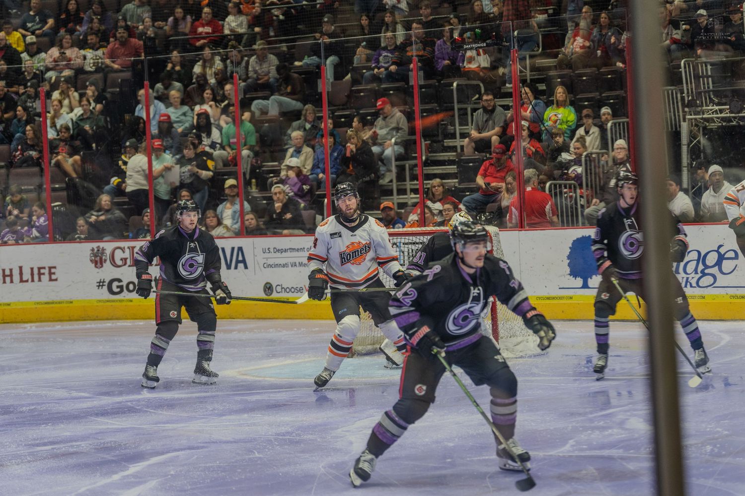 Multiple ice hockey players in purple and white jerseys compete for the puck during an indoor hockey game.