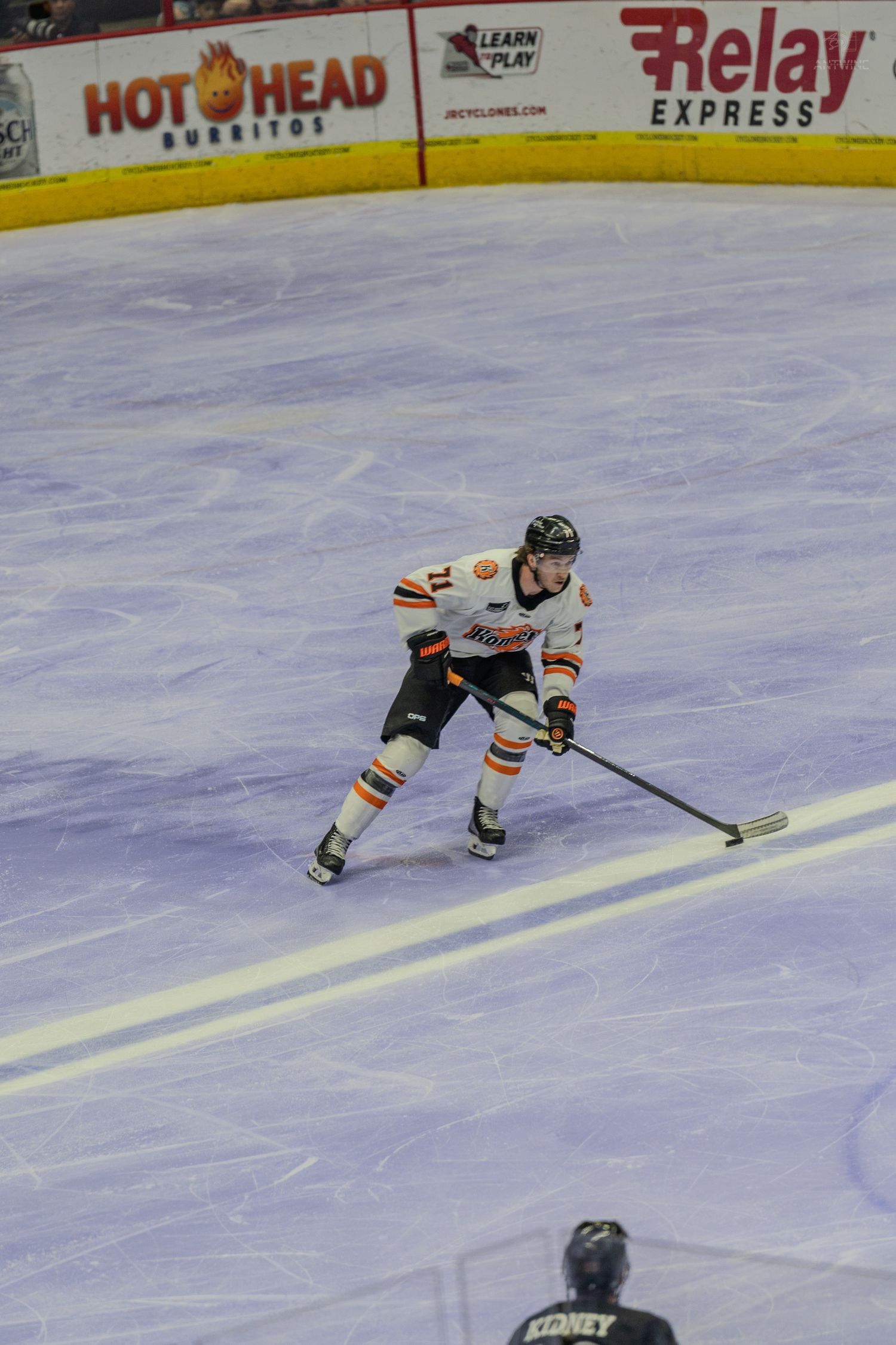 Hockey player in orange and white uniform handles the puck along the ice during a minor league game.