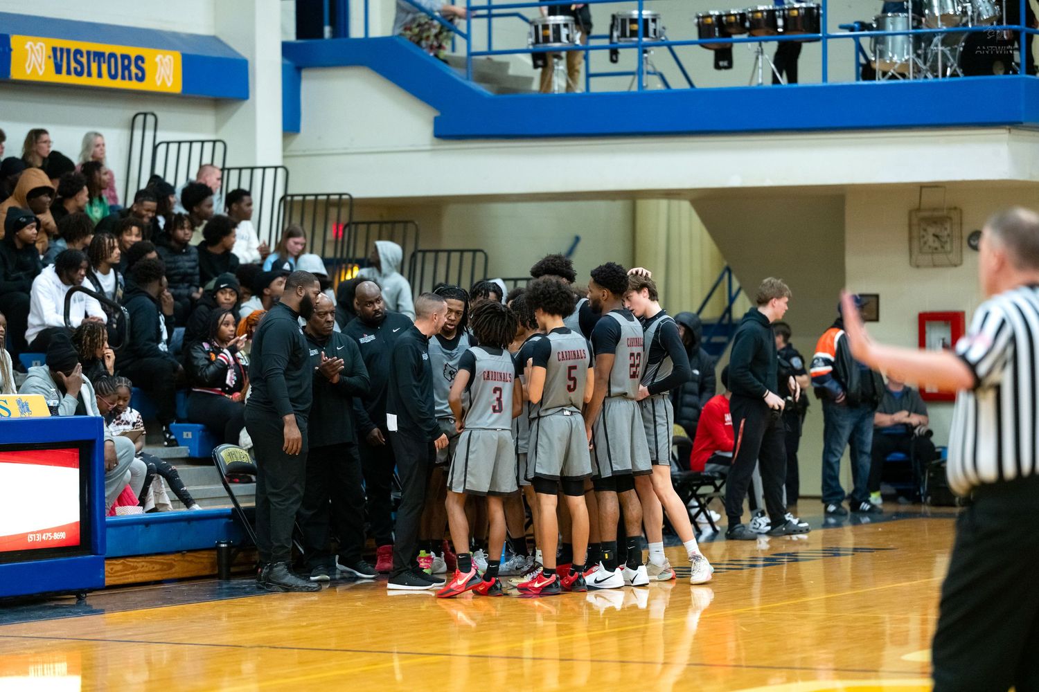 Visiting basketball team in gray uniforms huddles courtside during timeout in blue and white gymnasium.