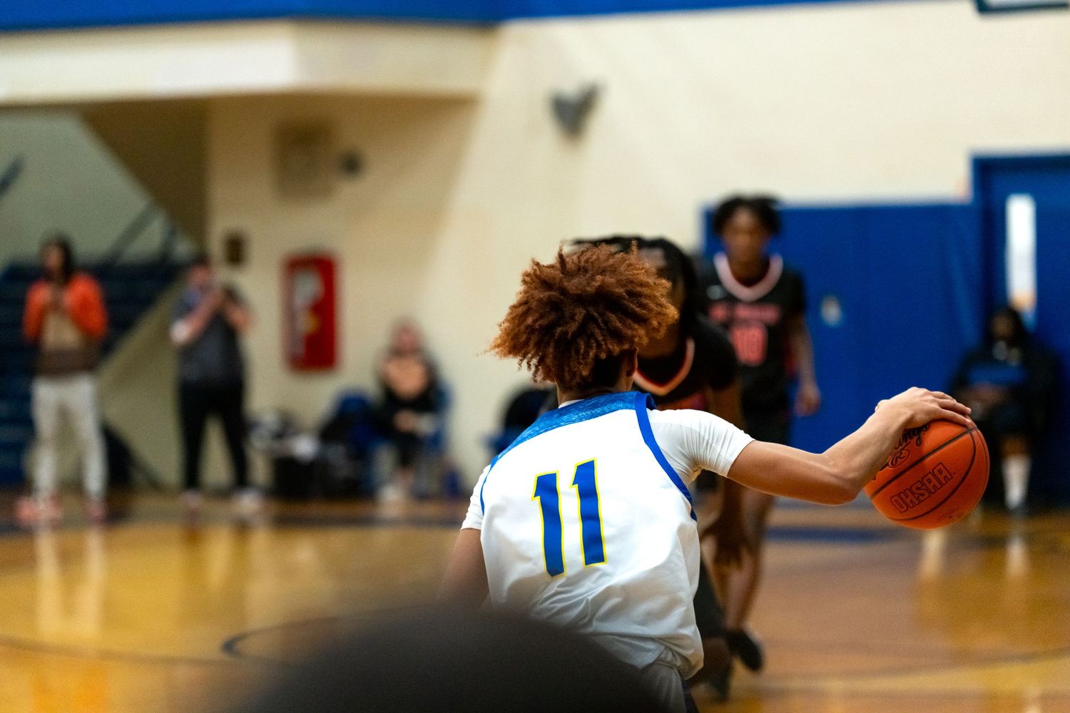 Player number 11 in a white jersey dribbles the basketball during a high school indoor basketball game.