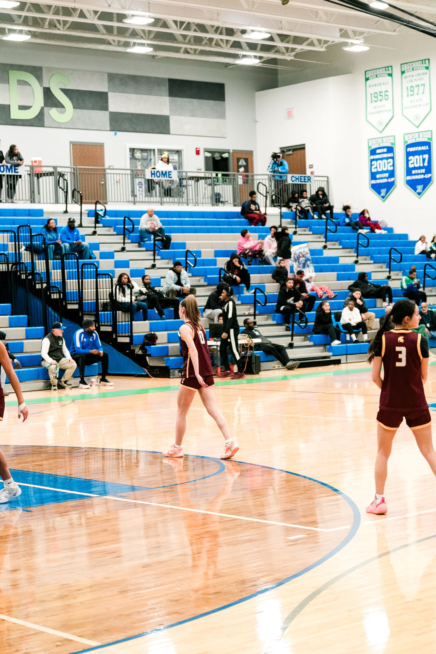 Female high school basketball players in maroon uniforms warm up on a blue and wood gymnasium court with sparse crowd.