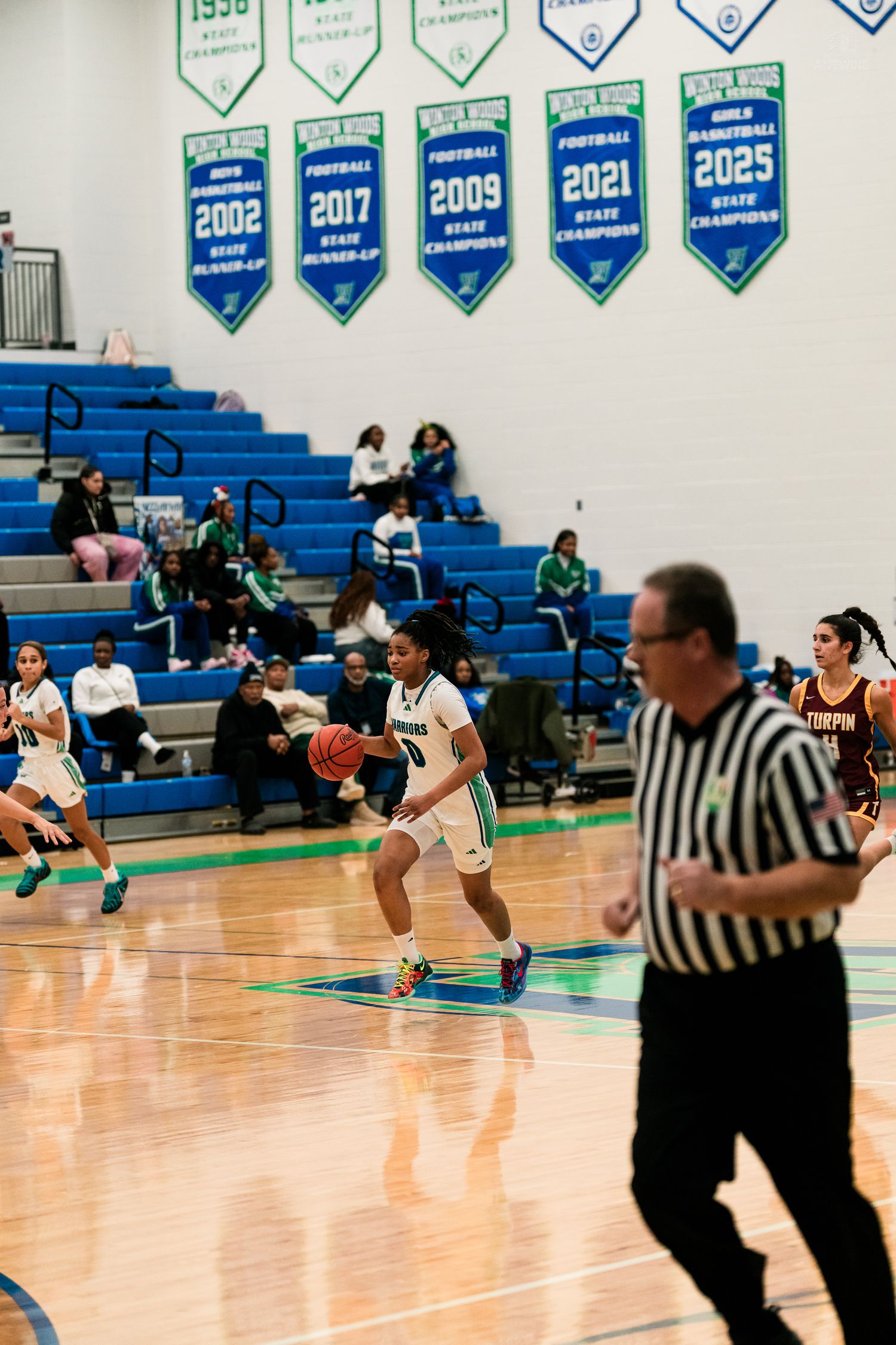 A girls high school basketball player in green dribbles past a defender with championship banners in background.