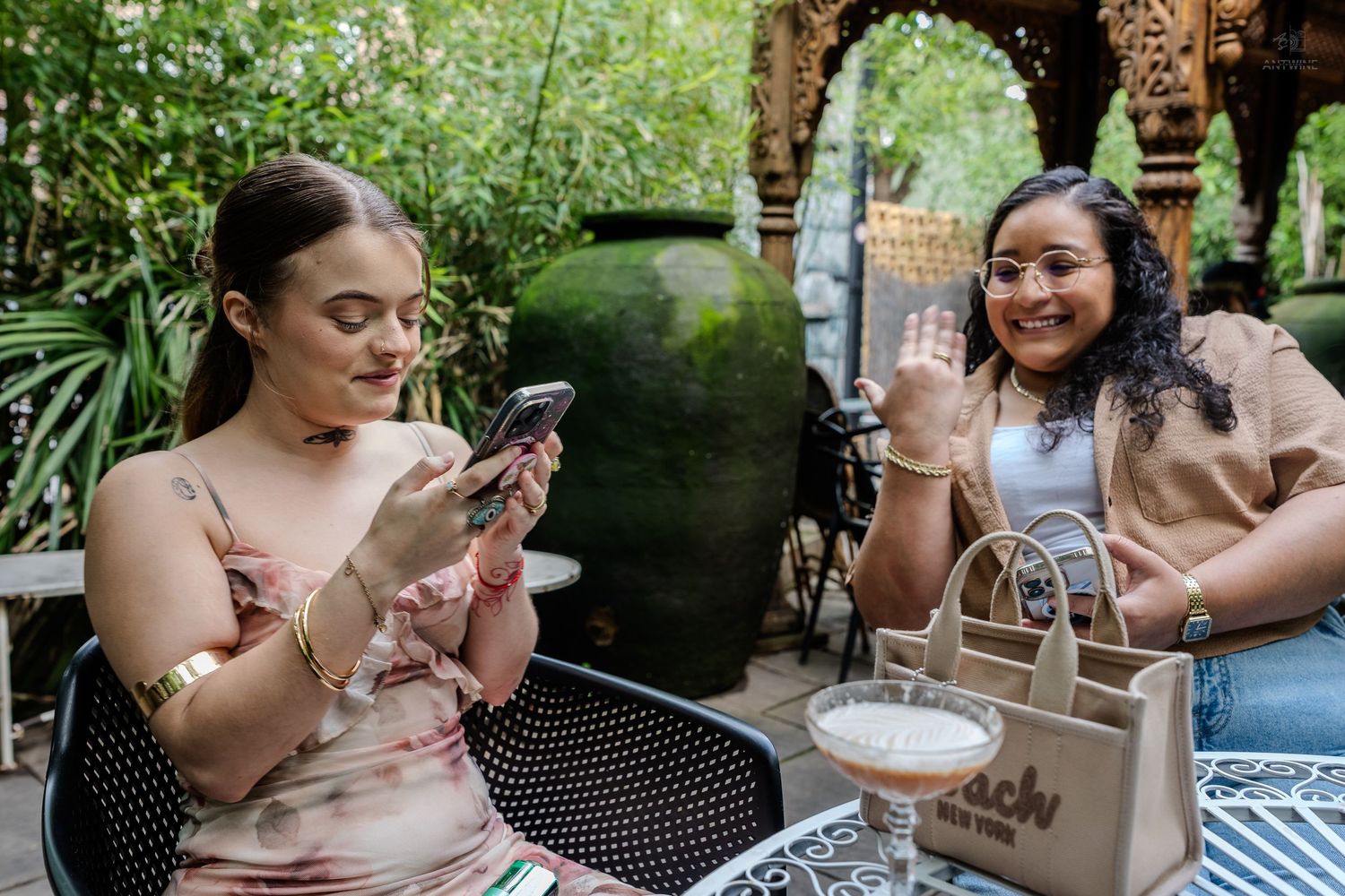 Two women at outdoor cafe table, one waving and smiling with cocktail and tote bag.