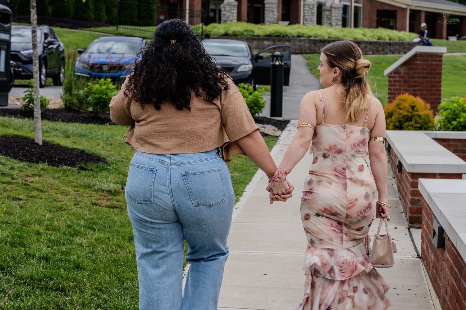 Two women holding hands walking down a sidewalk, one in jeans and one in a floral dress.