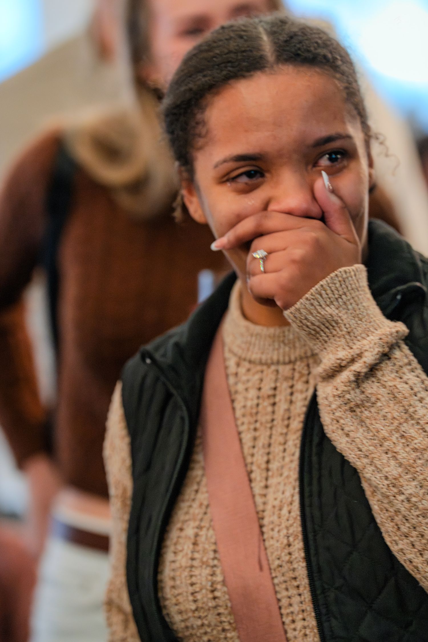 A young woman with natural hair covers her mouth while crying tears of joy at an indoor social gathering.