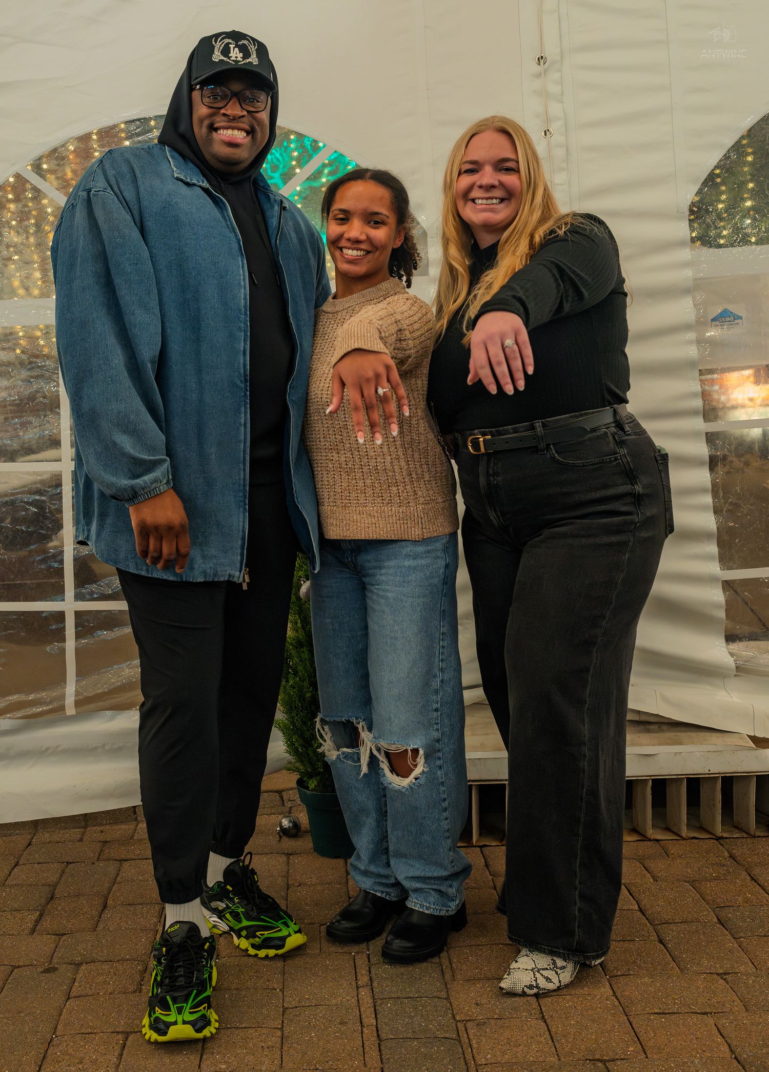 Three people smiling and posing together inside a white tent, woman in center showing engagement ring.