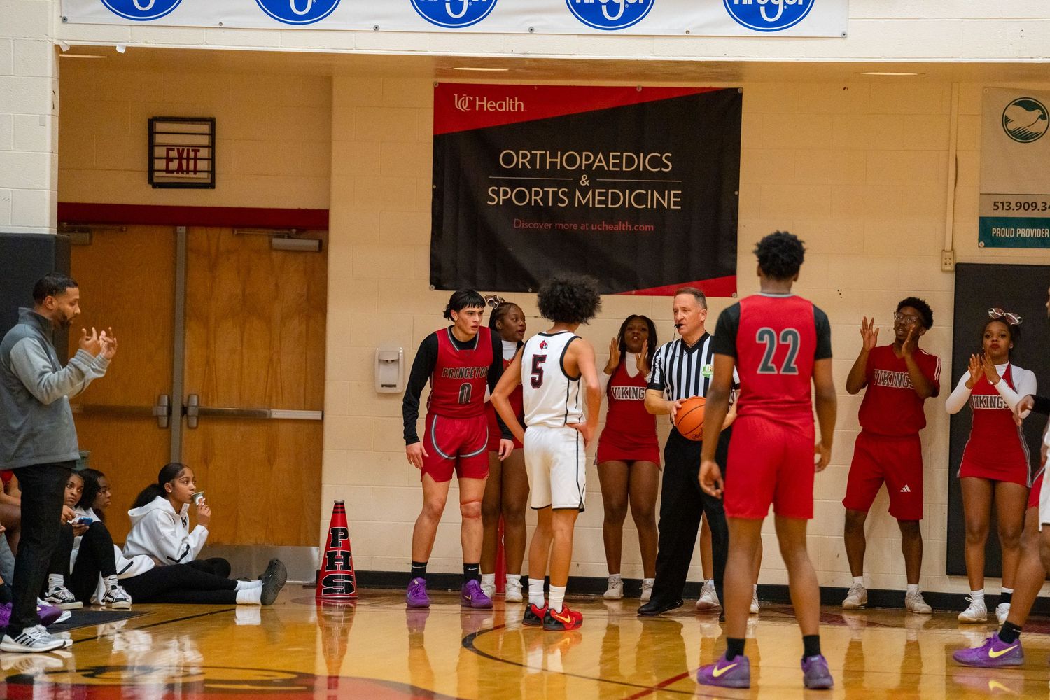 High school basketball players and referee gather at center court in a gym with an Orthopaedics Sports Medicine banner.
