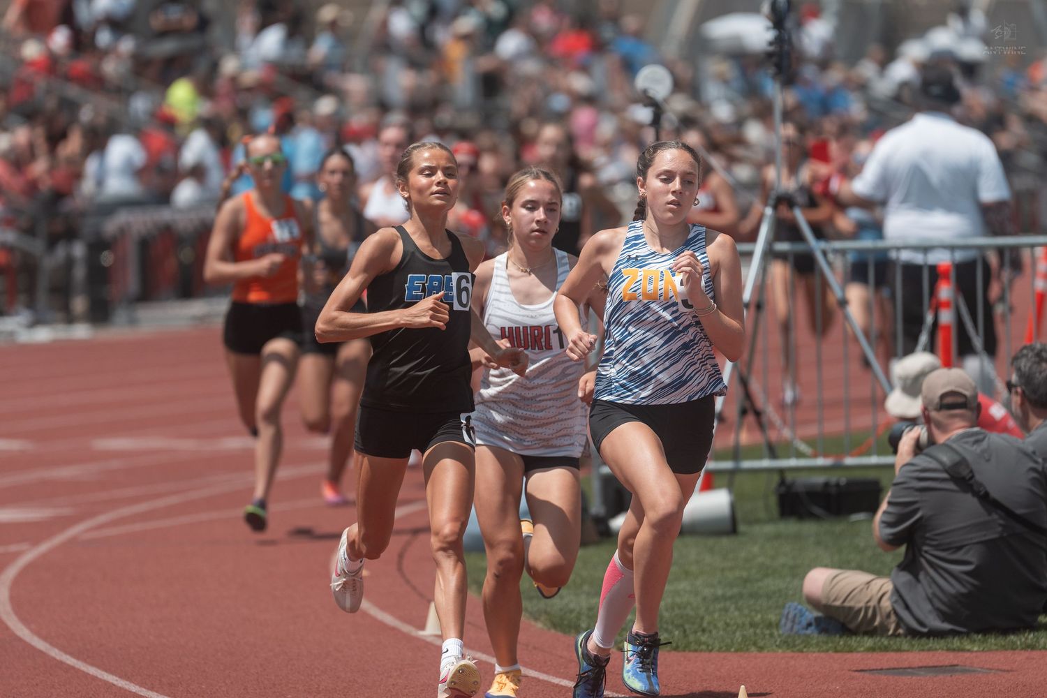 Three female runners compete closely together in a distance race at a sunny outdoor high school track meet.
