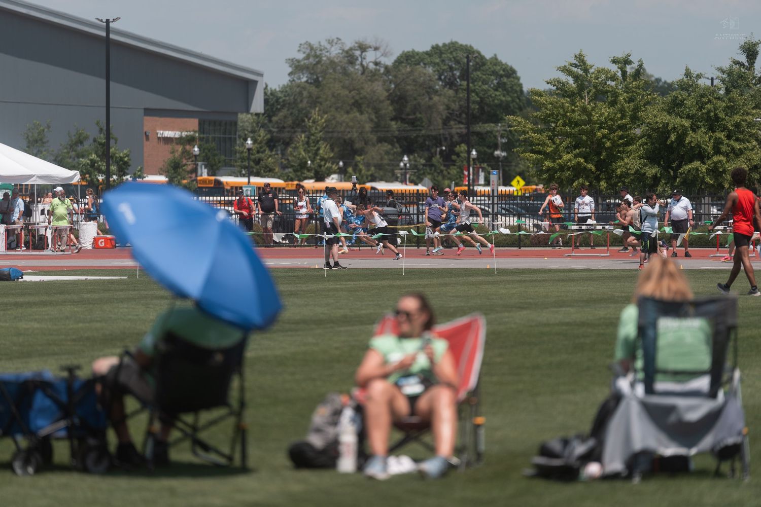 Spectators relax in lawn chairs under colorful umbrellas watching athletes compete at an outdoor track and field meet.