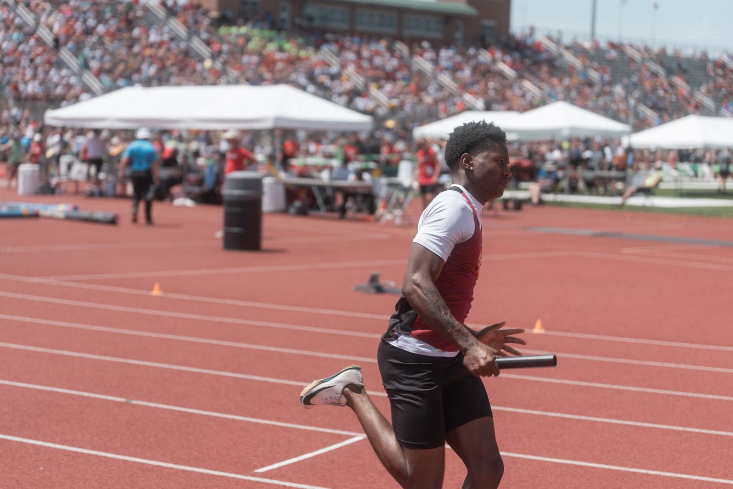 A sprinter carries a relay baton while running at a large outdoor track and field competition.