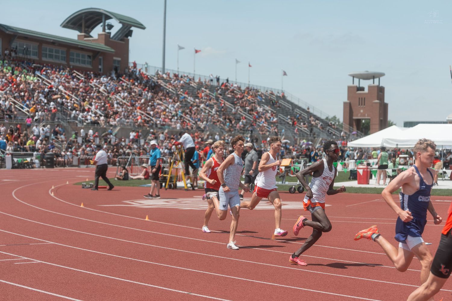 Athletes race on a red track at a crowded outdoor high school track and field competition on a sunny day.
